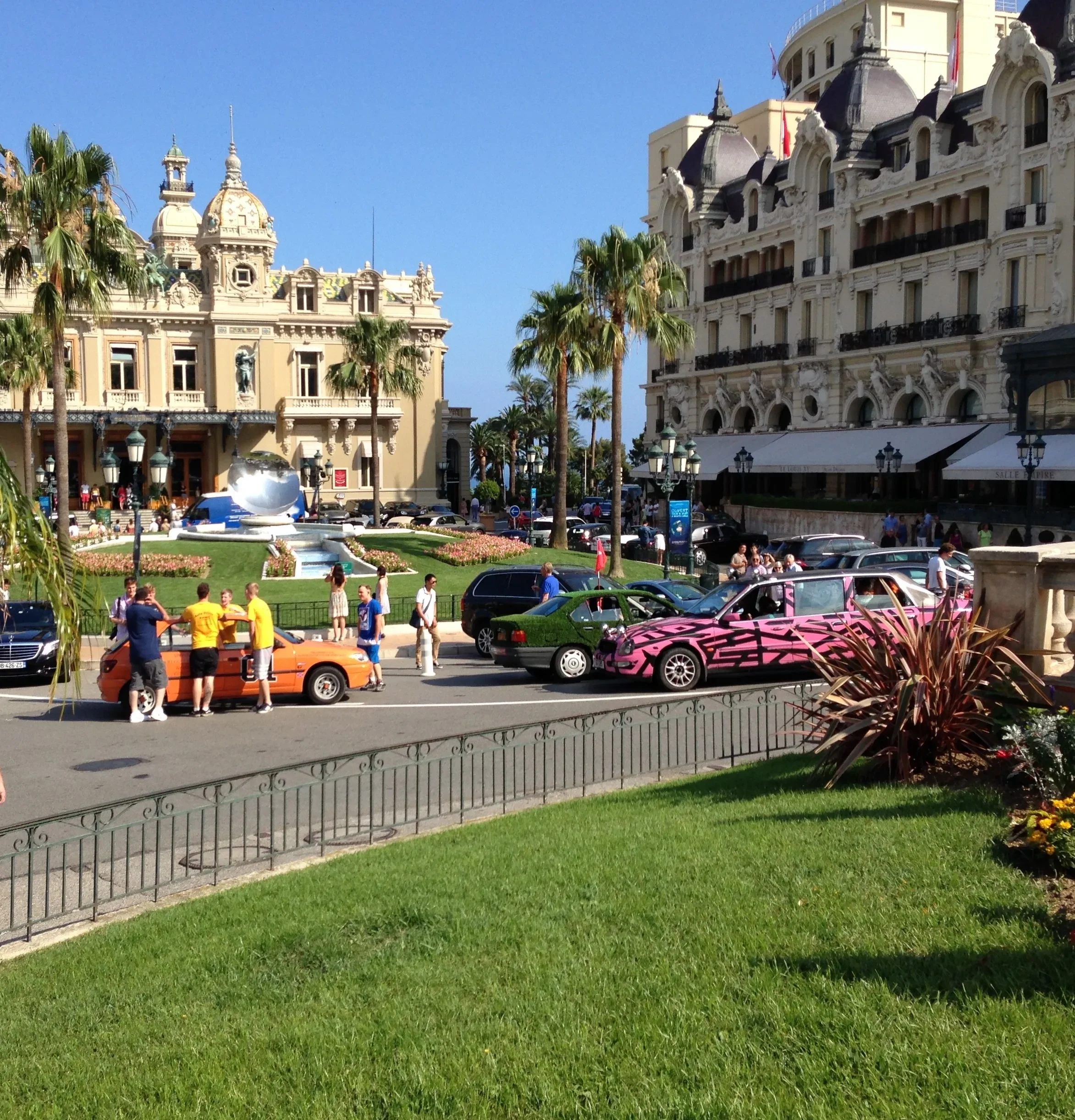Street scene with colorful cars, people walking, grand historic buildings, palm trees, and a park with flowers in a sunny, urban setting.