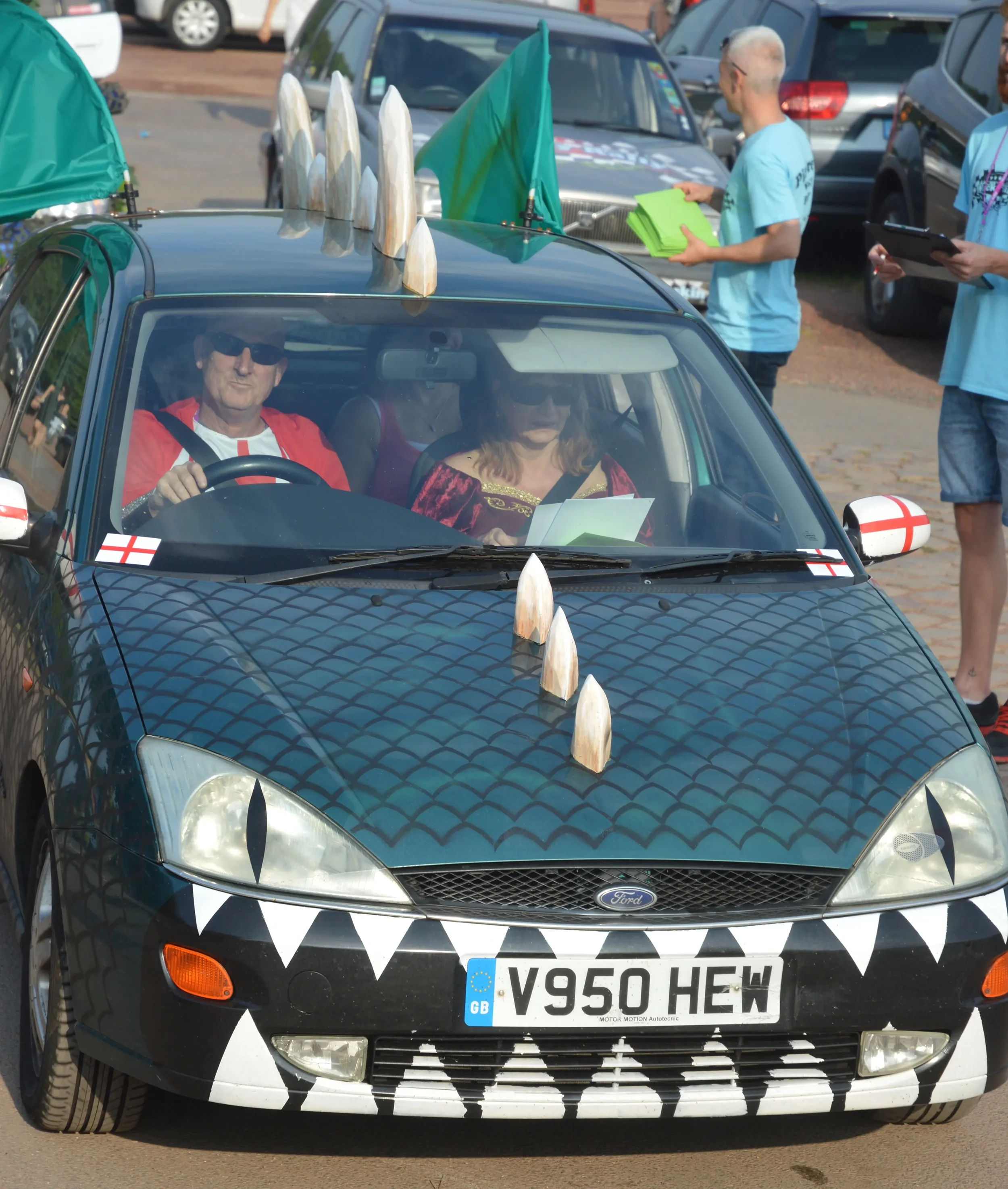 A black Ford car decorated with white and black geometric and flag-themed stickers, featuring a row of conical objects on the roof and a face design on the front bumper. Inside the car, two people are visible wearing sunglasses, and outside, people a