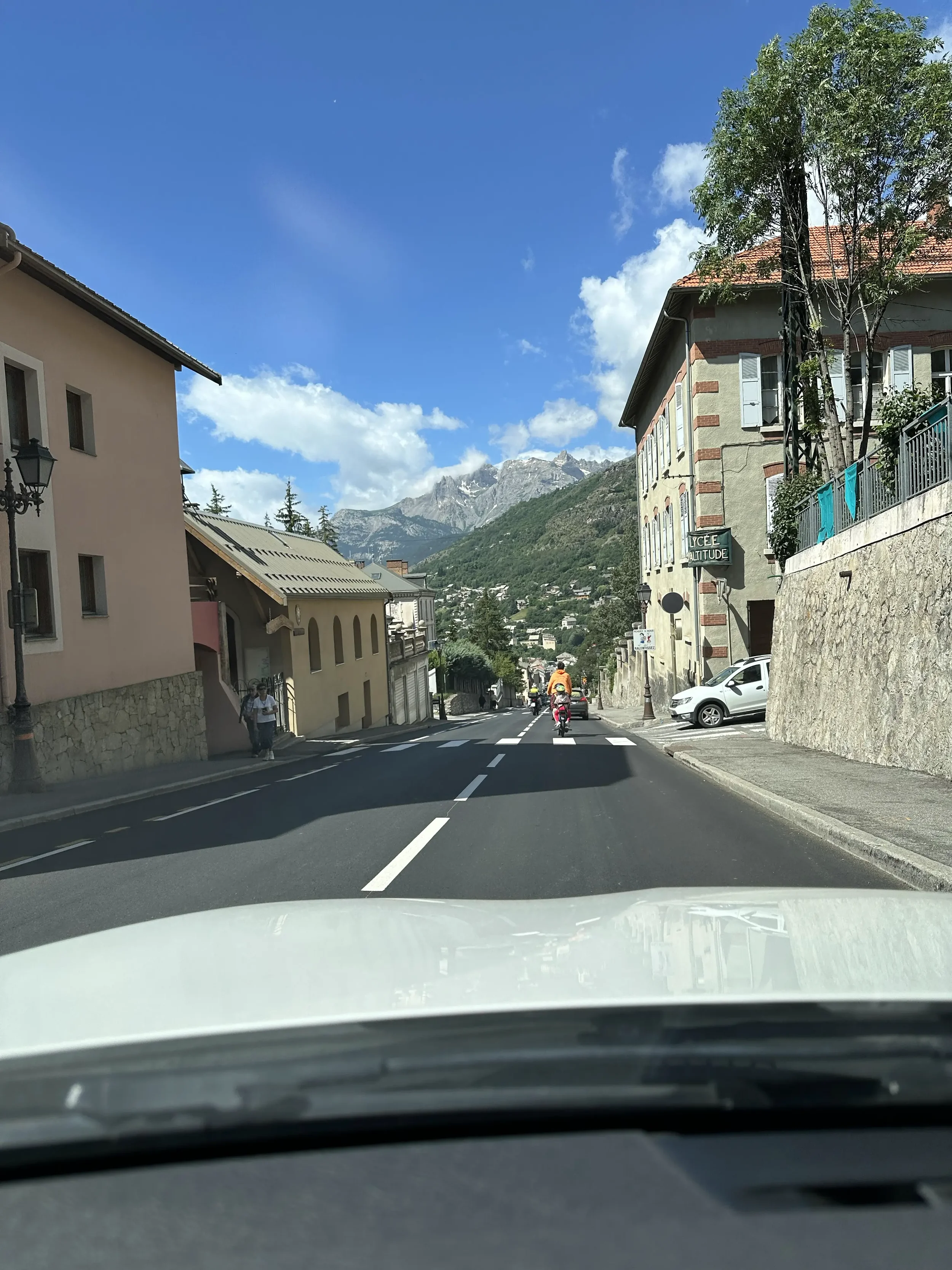 View from inside a car looking down a small mountain town street with buildings on both sides, a mountain in the background, partly cloudy sky, and a few pedestrians and cyclists on the road.