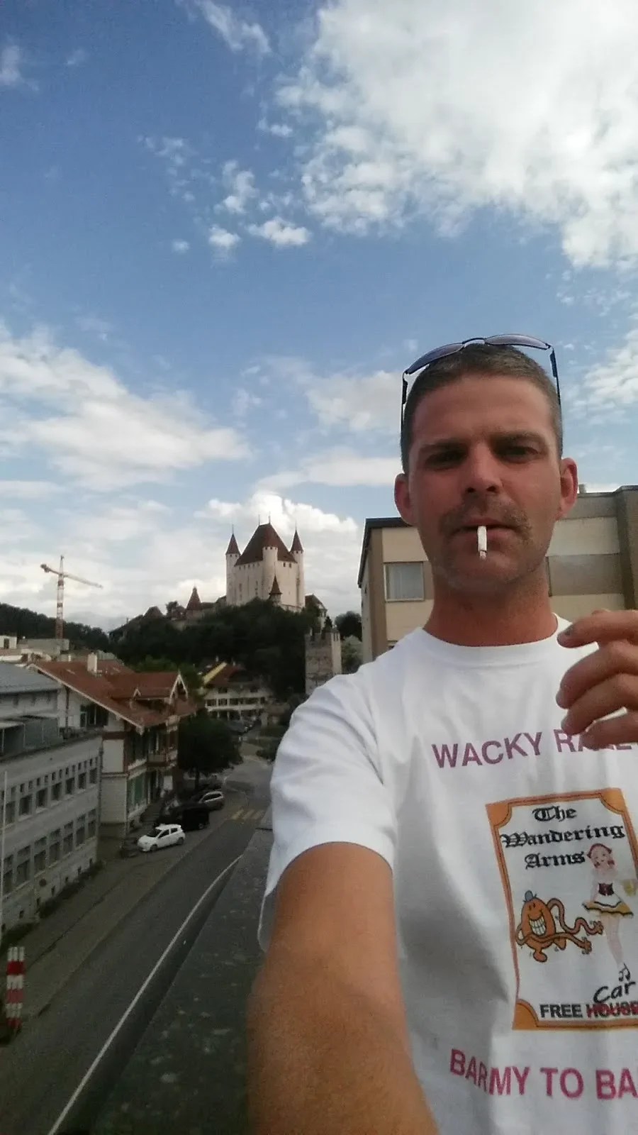 A man taking a selfie on a balcony with a castle in the background, holding a cigarette, wearing sunglasses on his head, and a white T-shirt with some text and a cartoon character on it.