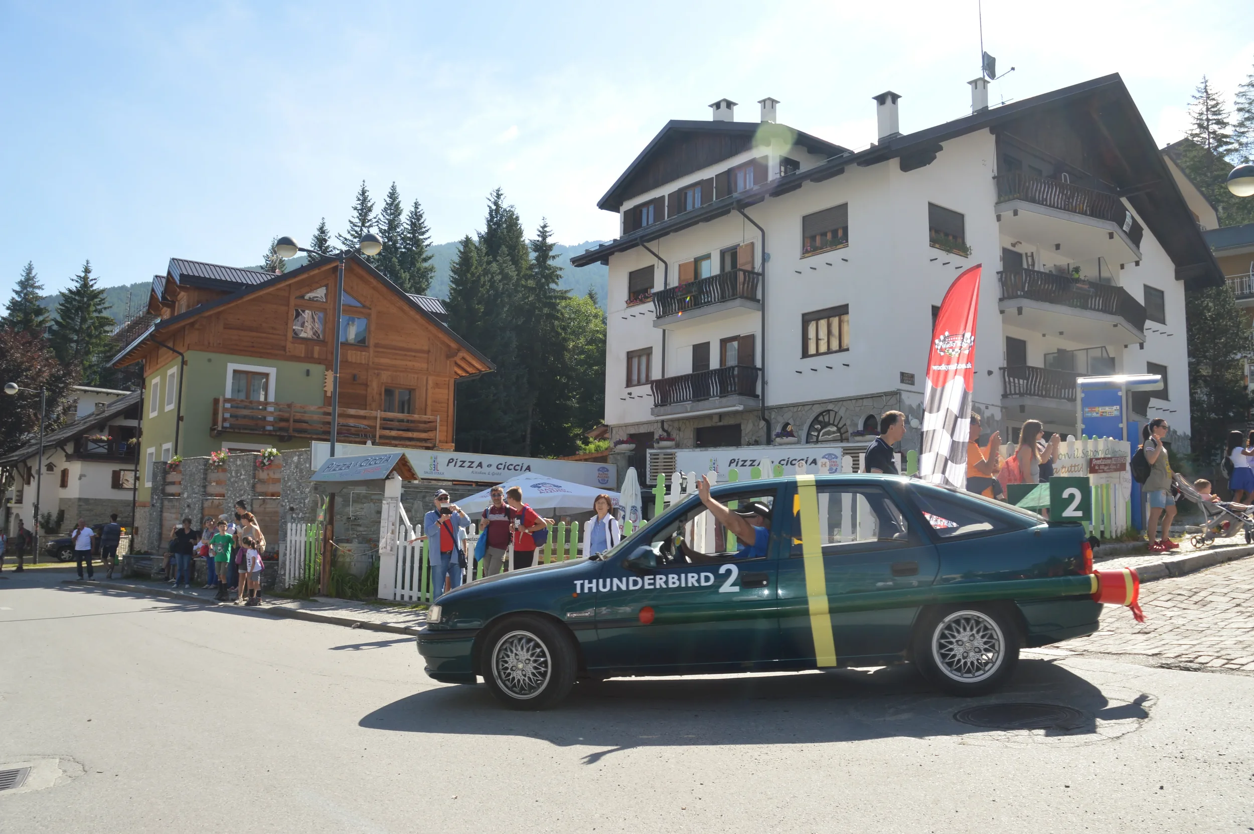 A parade scene in a mountain town with a fake car labeled "Thunderbird 2" driven by a person waving to the crowd. People line up along the sidewalk, with some taking photos. The buildings are chalet-style, with forested mountains in the background.
