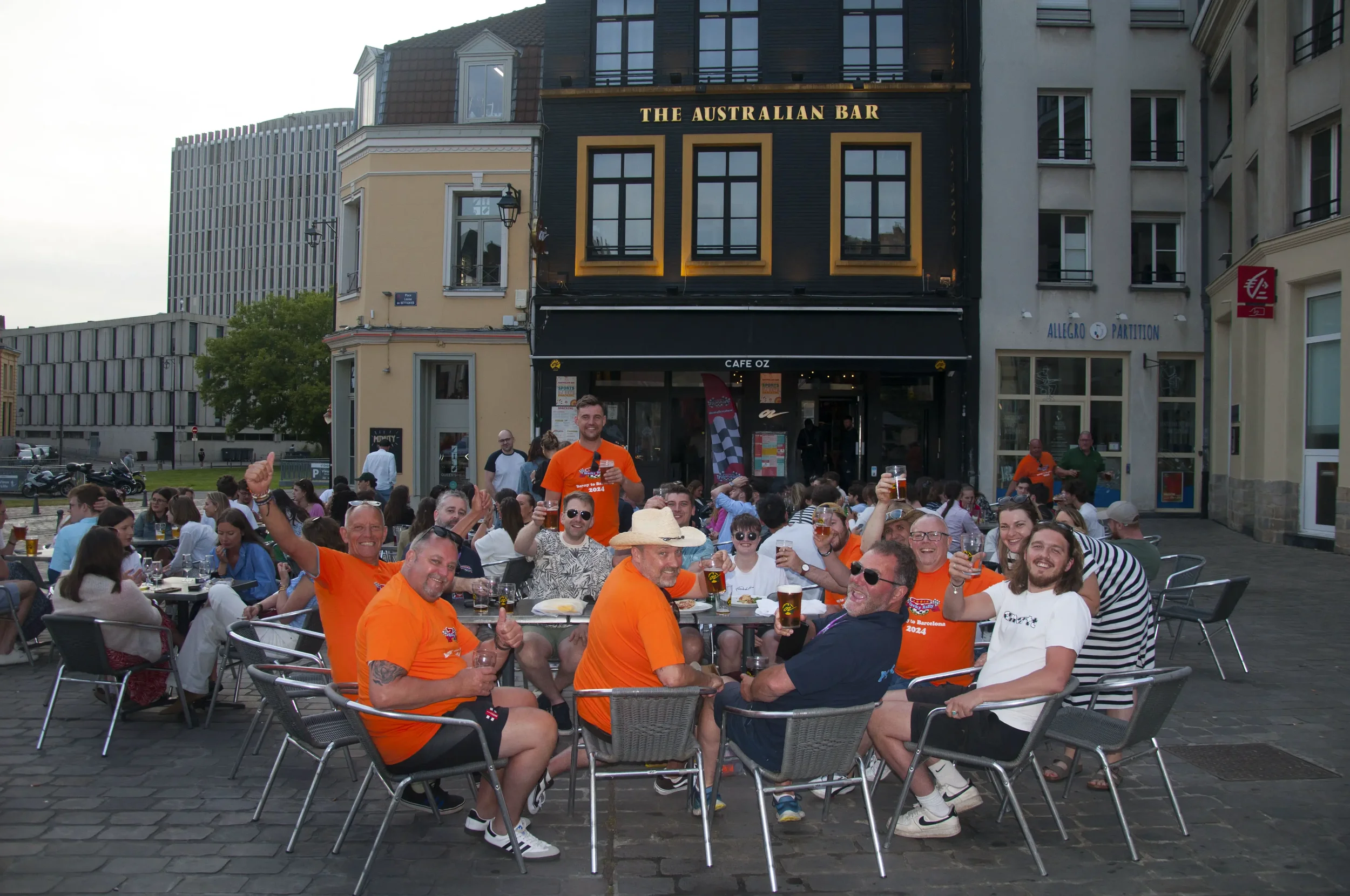 A group of people sitting and standing around outdoor tables at a bar, smiling and raising glasses in a toast. The bar is called "The Australian Bar," and the scene is lively with many patrons, some wearing orange shirts. It is an evening setting wit