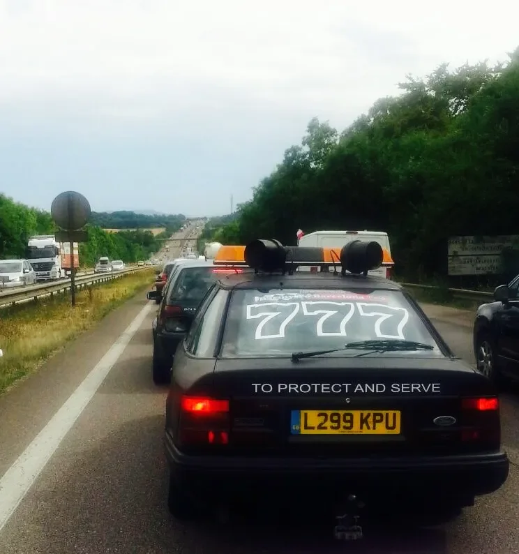 Traffic jam on a highway with several cars and trucks, with a black car in the foreground displaying large white '777' numbers on the back windshield and a slogan 'TO PROTECT AND SERVE' on the rear.