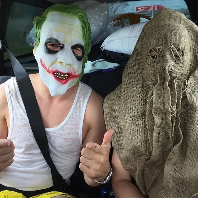Person wearing the Joker mask from Batman and a sleeveless shirt, giving two thumbs up, sitting in a car with cluttered items including a burlap sack with eyes stitched on it.