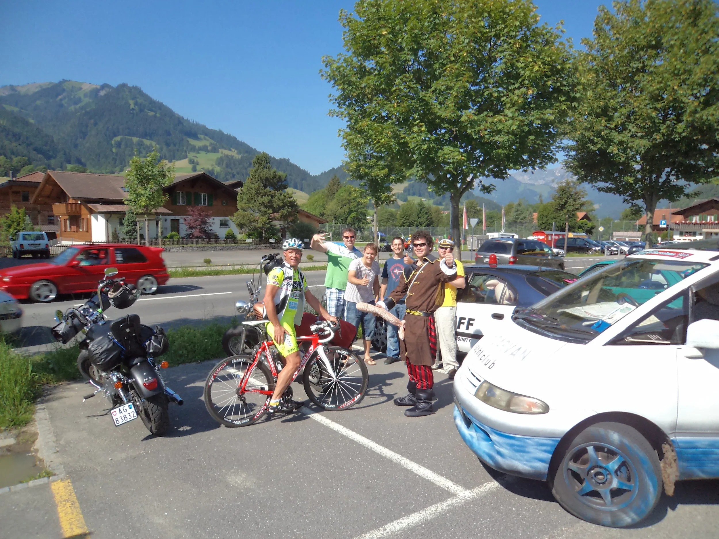A group of people, including a cyclist in a bright yellow outfit, gathered in a parking lot with a mountainous landscape in the background. Some are giving thumbs-up, and a man dressed in a pirate costume is pointing at the camera. Several motorcycle
