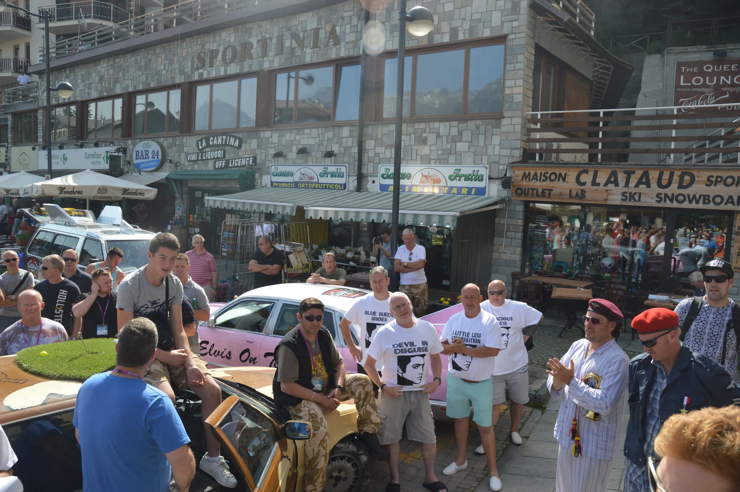 Crowd gathered outdoors in front of shops and restaurants, with some people sitting on a car and others standing, engaged in conversation or observing. Buildings have signs for various businesses like sports shops, a bar, and a grocery store. The bac