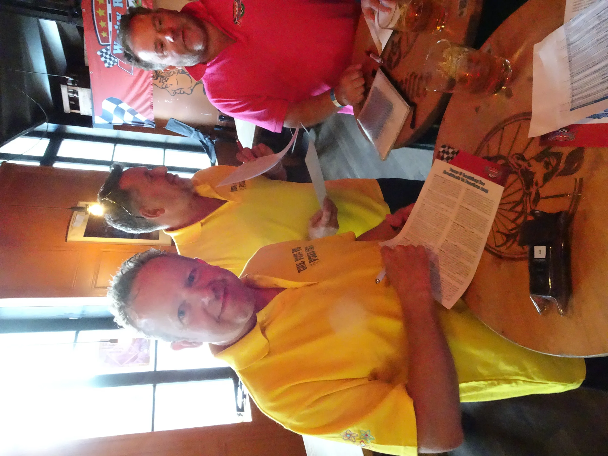 Two men in yellow shirts and one man wearing a red shirt are standing and sitting at a wooden table in a restaurant. They are reading menus. The table has glasses of drinks, a calculator, and papers on it. The background has a window and a wall with 