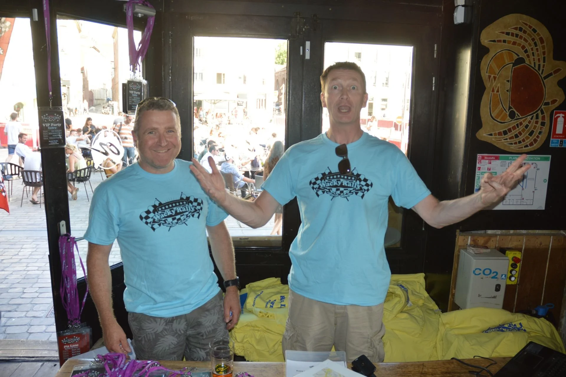 Two men standing inside a building, wearing matching light blue t-shirts with a checkered flag design and the words "Wacky Rally" on them. One man is smiling, and the other has his arms raised with a surprised expression. There is a table with pink l