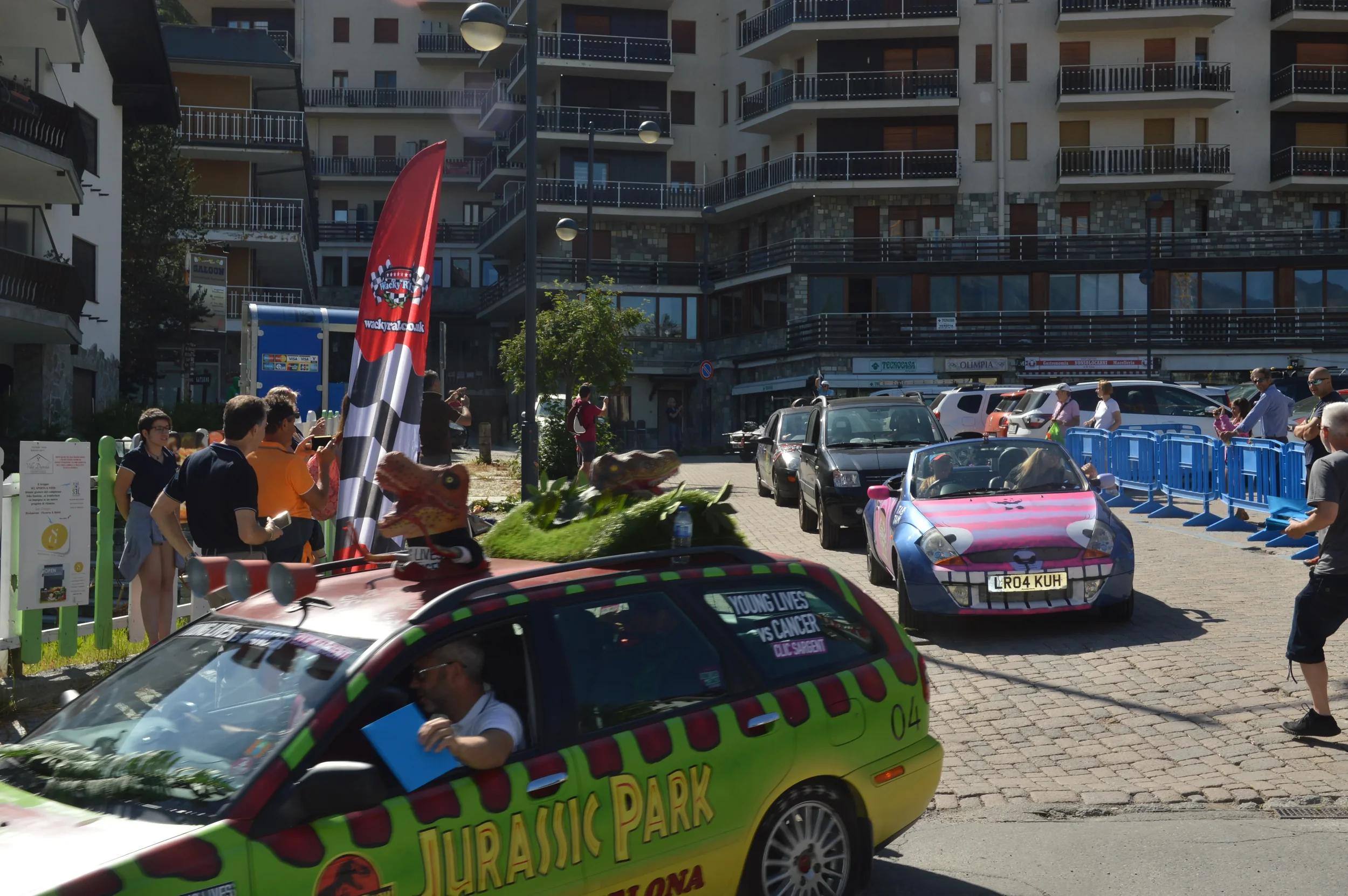 A gathering of people around a colorful car decorated with 'Jurassic Park' theme in an urban area, with cars parked and a tall residential building in the background, on a sunny day.