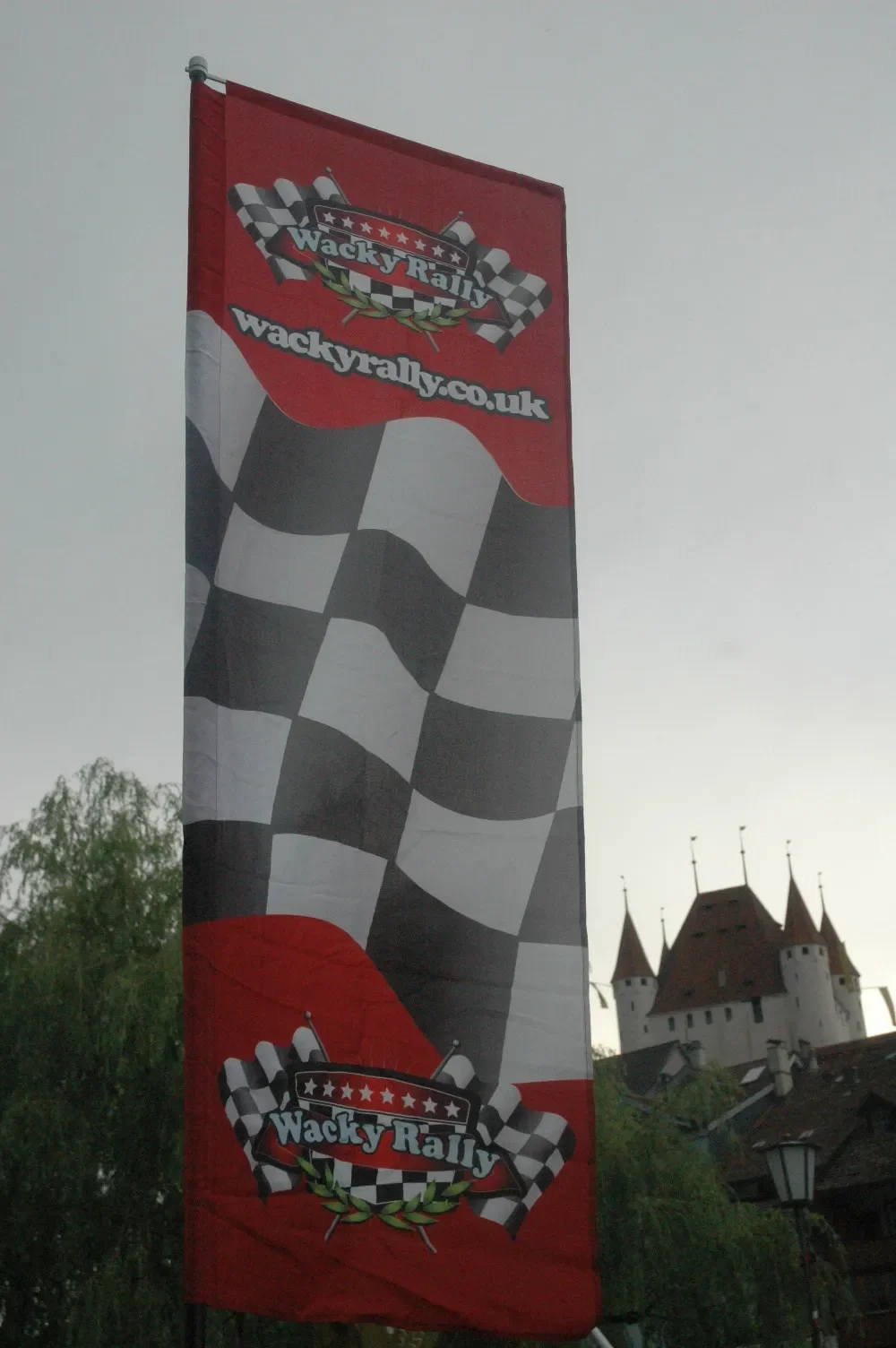A vertical flag with a checkered racing flag pattern and the logo "Wacky Rally" on a red background, flying outdoors near a castle-like building.
