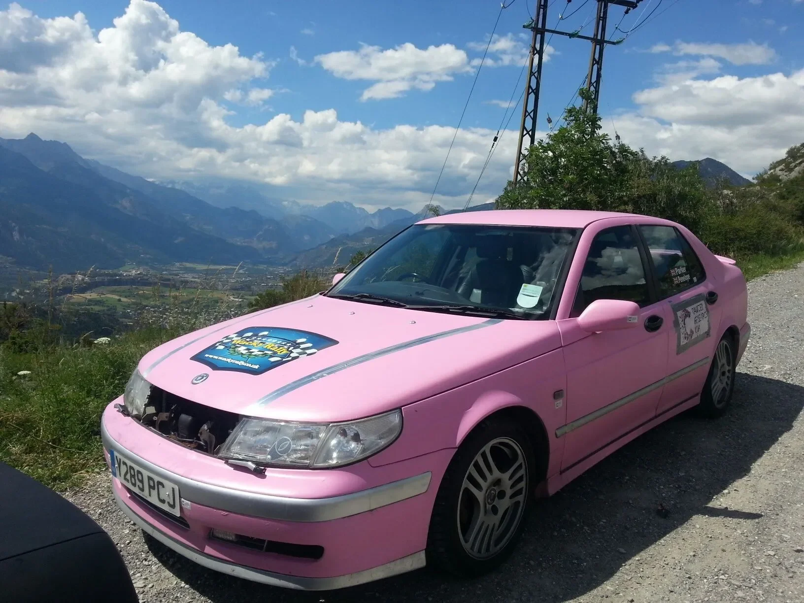 Pink car parked on a mountain road with mountains and a valley in the background.