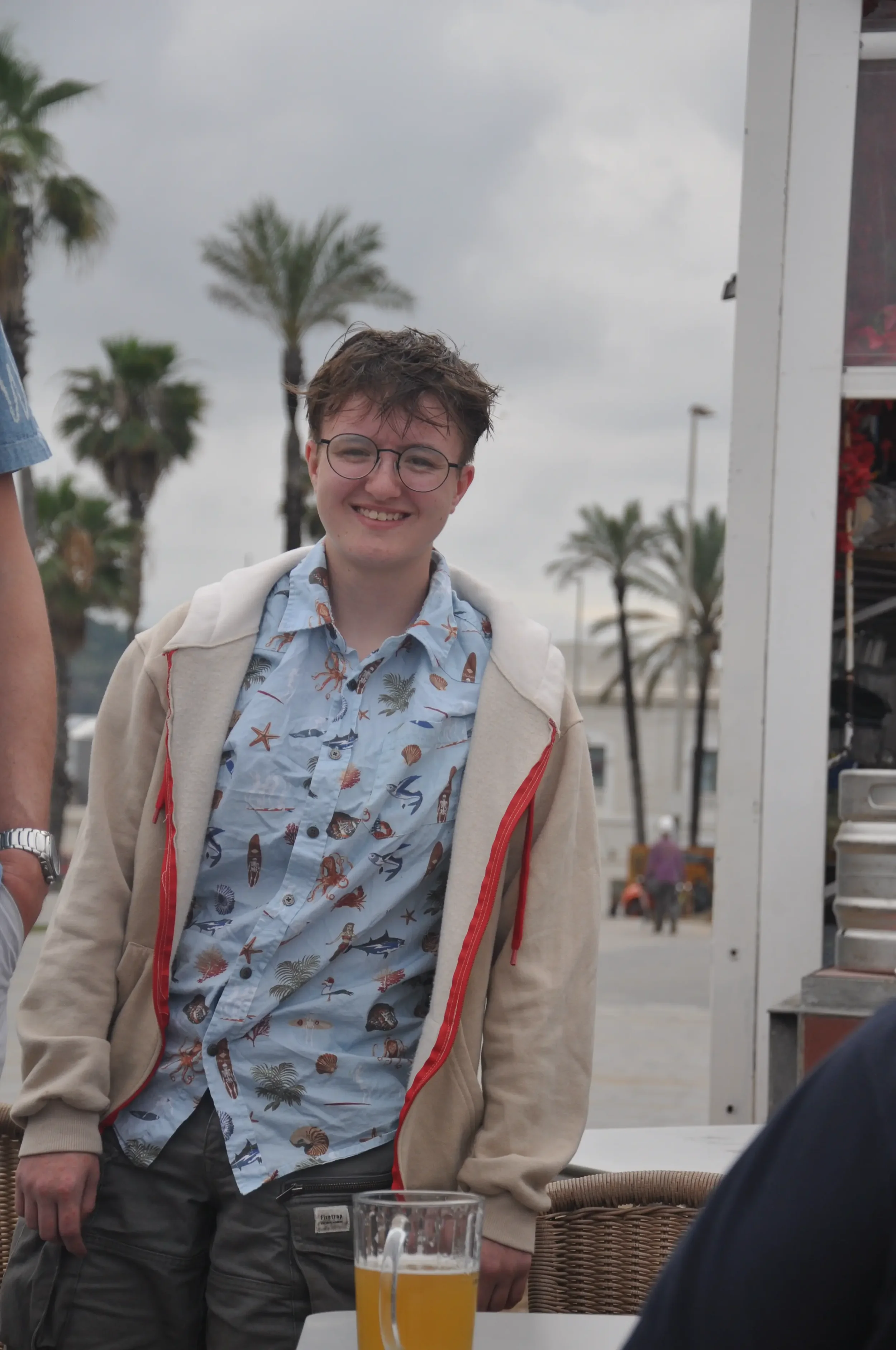 A young man with glasses, wearing a hawaiian shirt and a beige zip-up hoodie, standing outdoors with palm trees in the background. There is a glass of beer on the table in front of him.