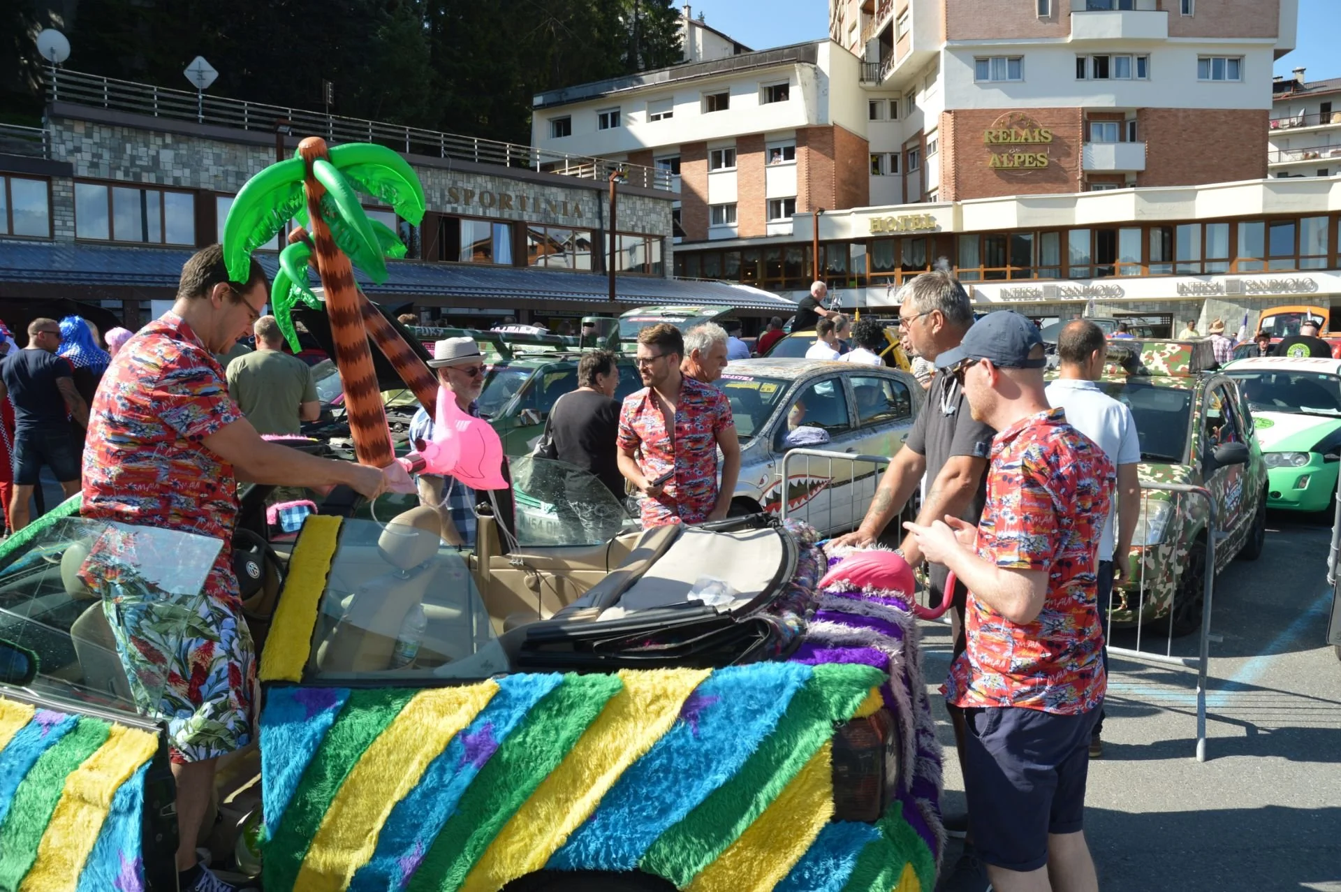 People at a car show with vintage cars decorated with colorful towels and accessories, some individuals wearing tropical shirts and inflatable flamingos.