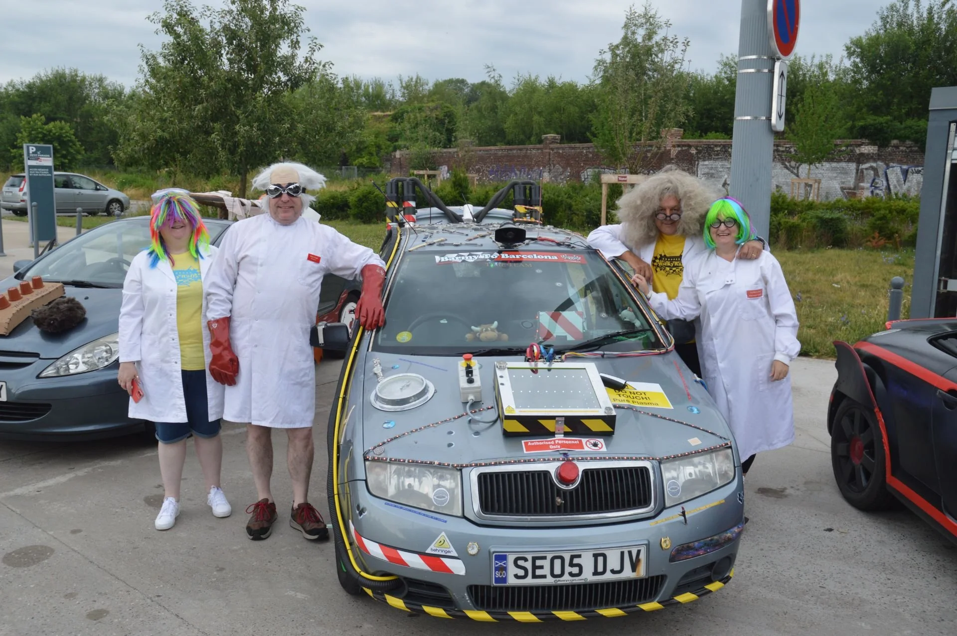 Four people dressed as scientists in humorous costumes standing next to a modified car with various gadgets and signs.