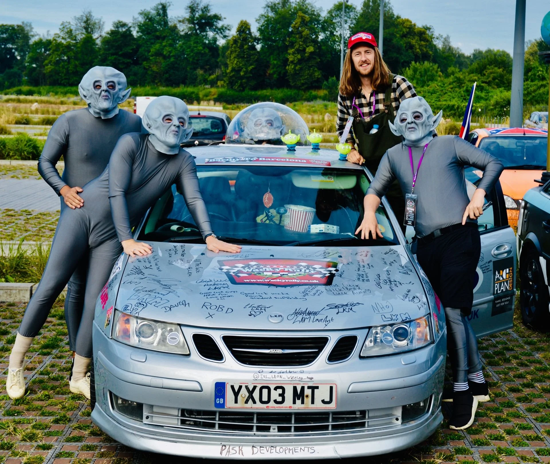 Four people dressed in alien costumes posing on a signed car at an outdoor event, with a person in casual clothing standing among them.