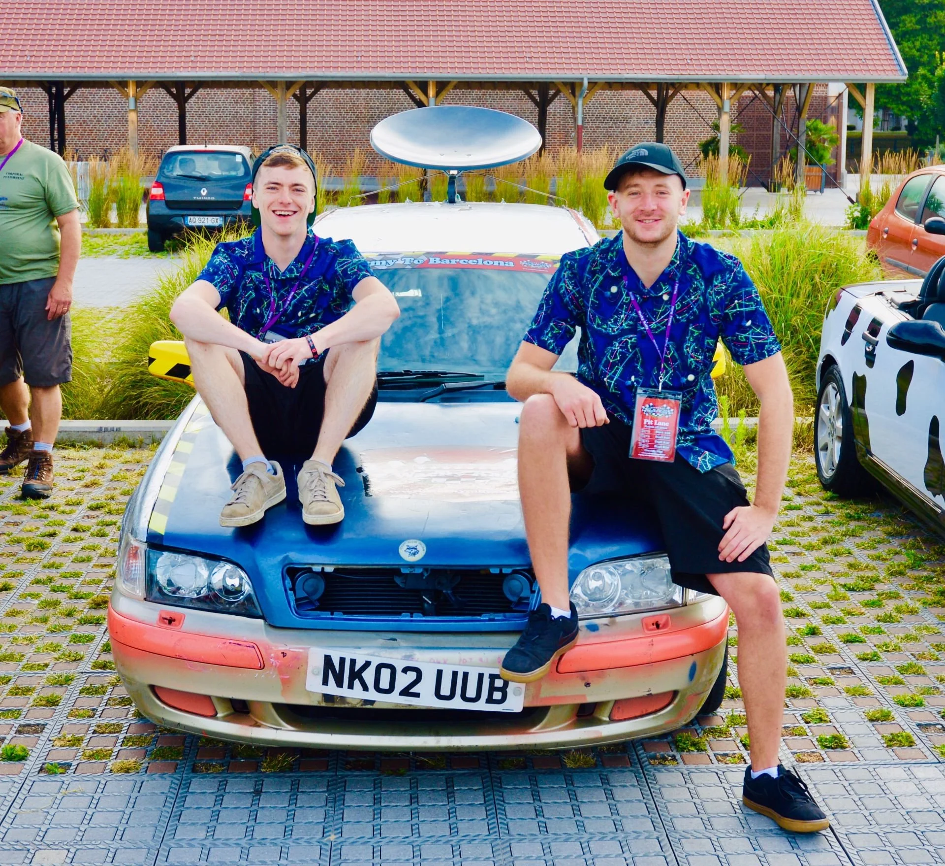 Two young men sitting on the hood of a vintage race car, smiling at the camera, at an outdoor event. One man has short hair and wears a blue patterned shirt and black shorts, the other has a hat and wears a similar shirt and shorts. There is a radio 