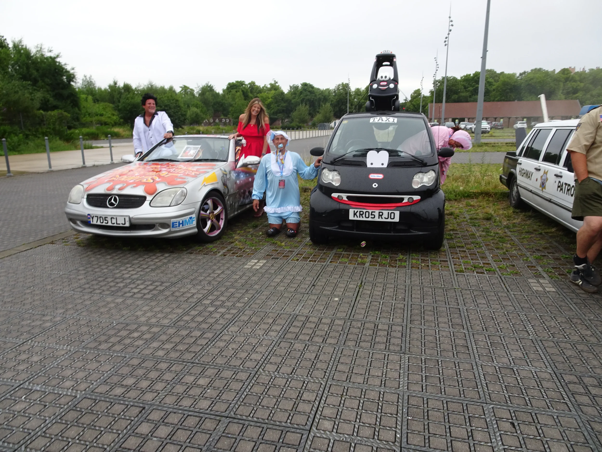 Group of people parked with decorated cars, including a Mercedes convertible with artwork and a small black vehicle with cartoon face decorations, in an outdoor lot with trees and cloudy sky.