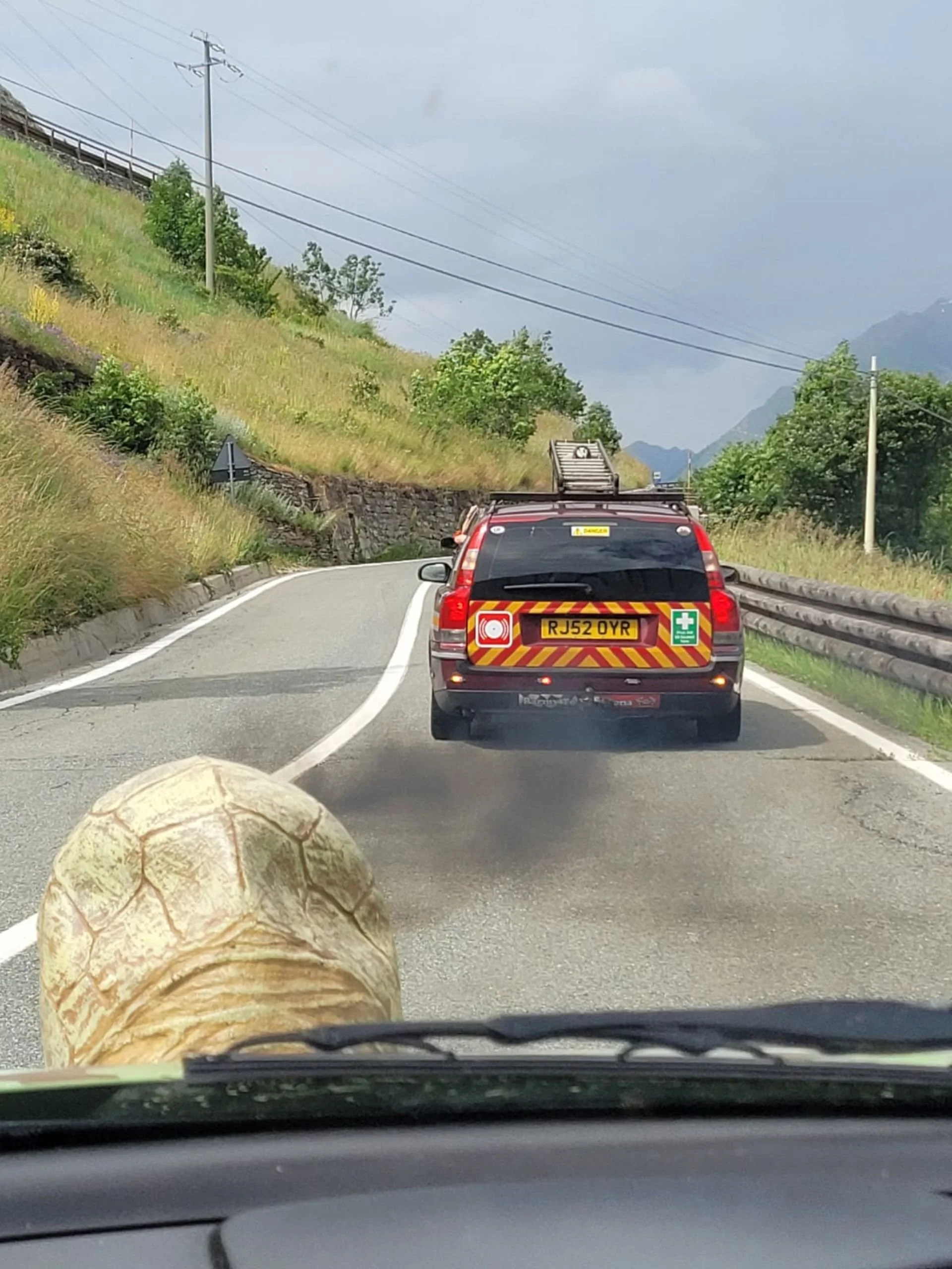 A Lamborghini Aventador with a fire truck behind it on a mountain road, with hills and power lines in the background.
