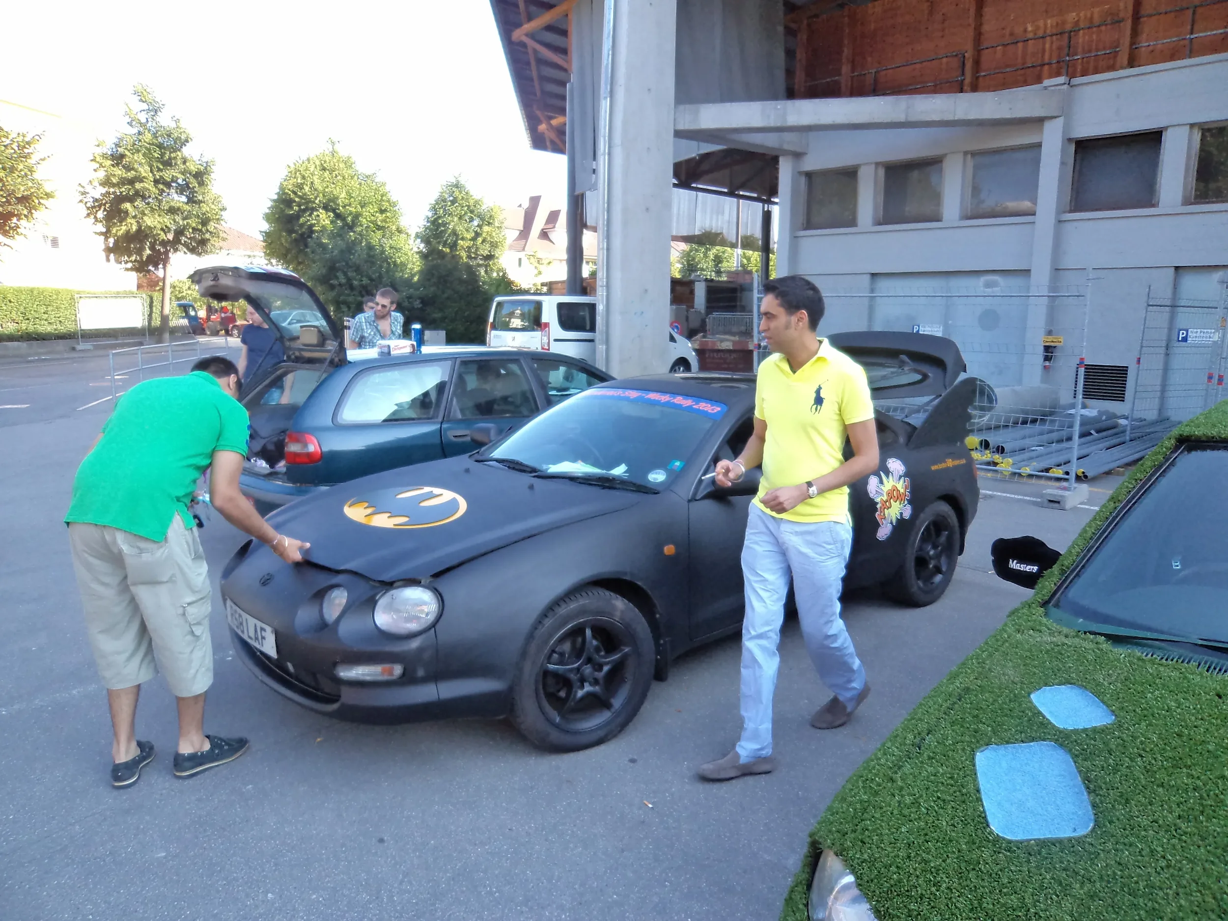 Two men working on a black car decorated with Batman logos in a parking lot, with one man inspecting the front and another walking nearby.