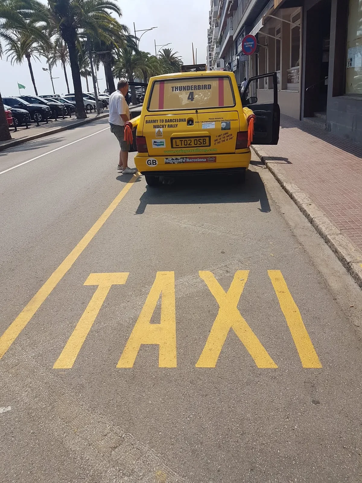 A yellow taxi parked on the side of a street with a man standing beside it. The taxi has various stickers and text, including 'TAXI' painted on the street in front of it and 'Thunderbird 4' on the back of the car.