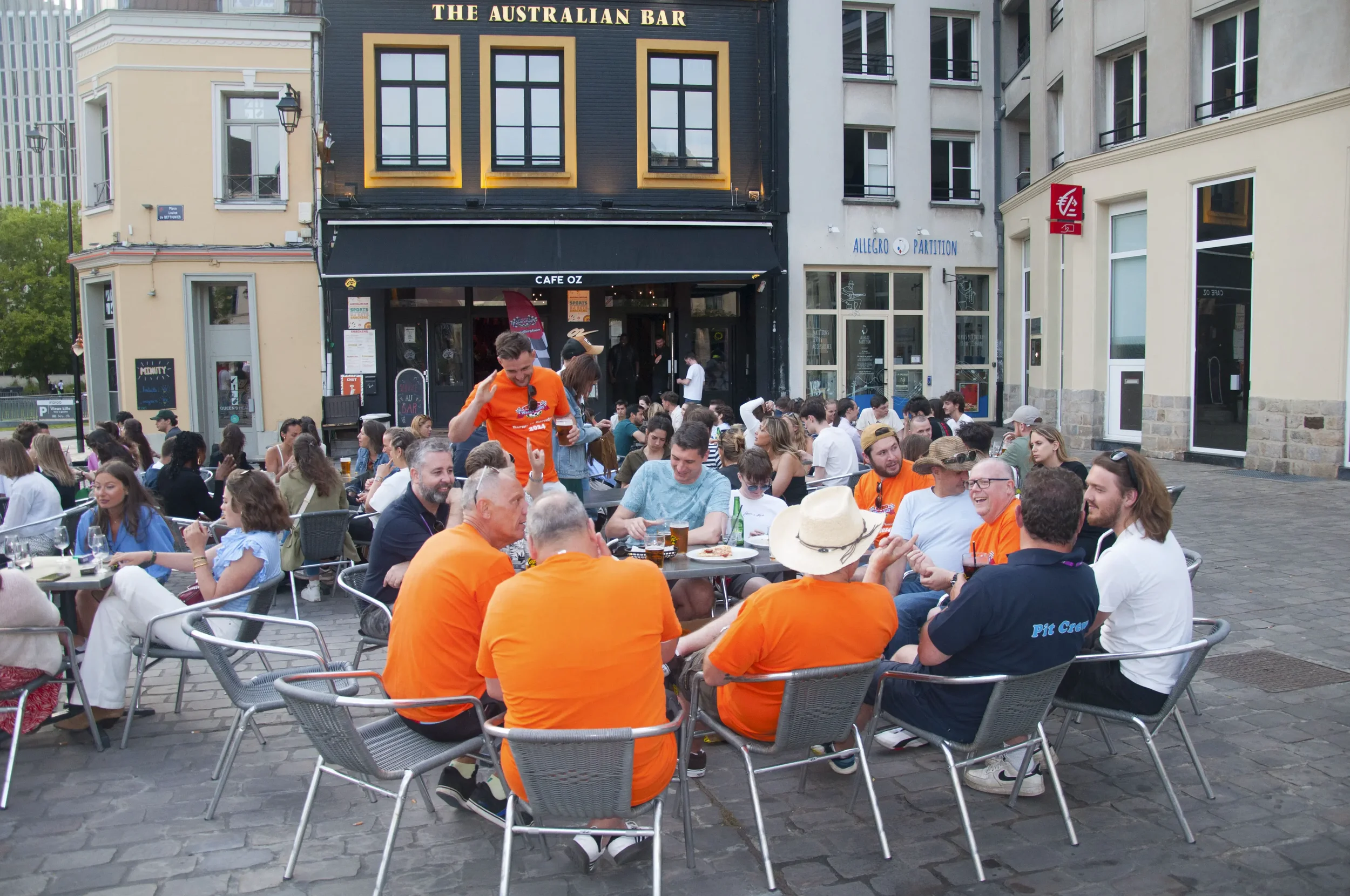 People sitting and socializing at an outdoor dining area in front of cafes and bars on a city street.