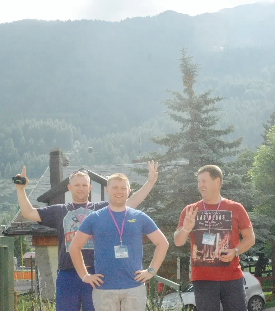 Three men smiling outdoors with mountains and trees in the background. The man on the left is making a peace sign with his right hand. The man in the middle has his hands on his hips, and the man on the right is waving with his left hand. All three a