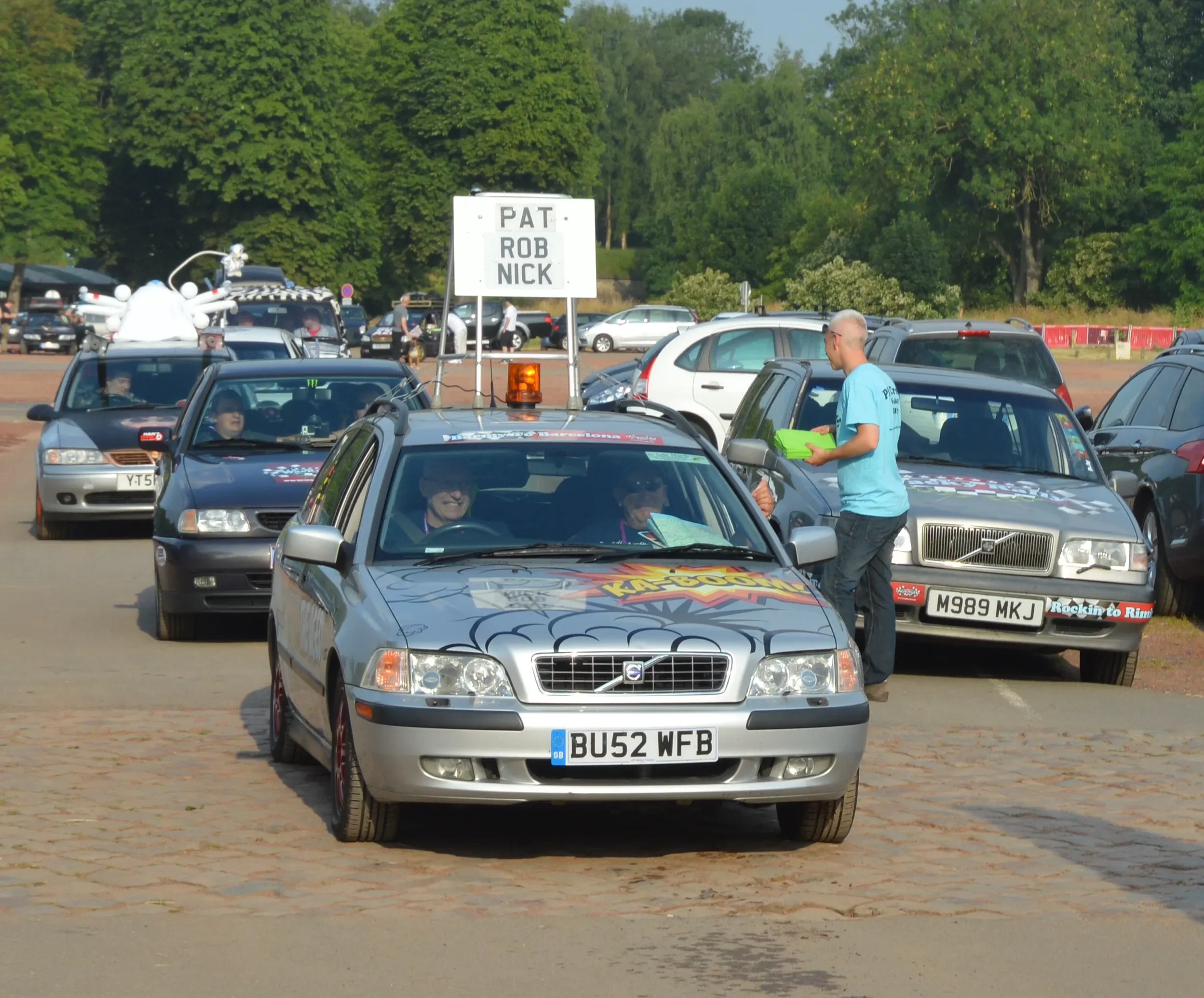 Line of cars in a parade, with a man in a turquoise shirt handing out flyers and a sign on one of the cars that reads 'PAT, ROB NICK', suggesting a rally or celebration event.