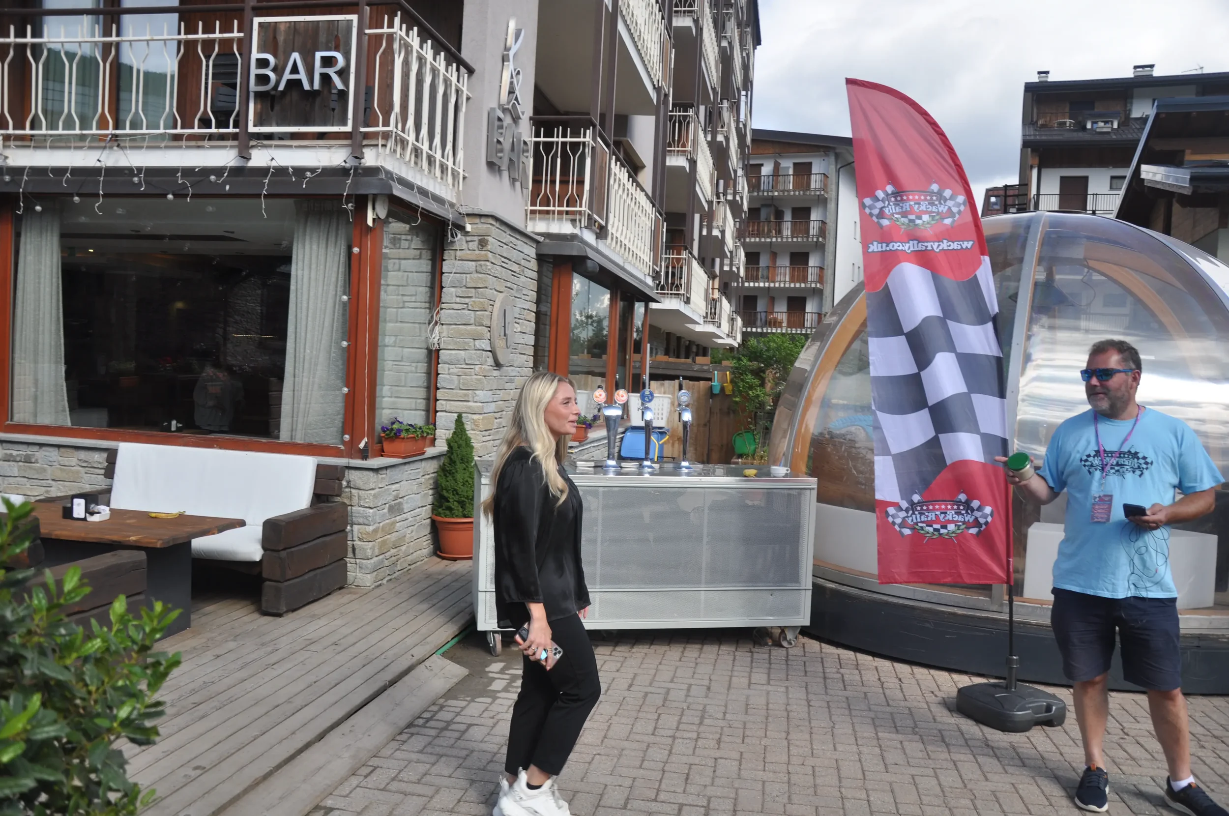 Two people standing outside a building with a bar sign, near a flag with checkered pattern, under an outdoor structure, with a beer tap station in the background