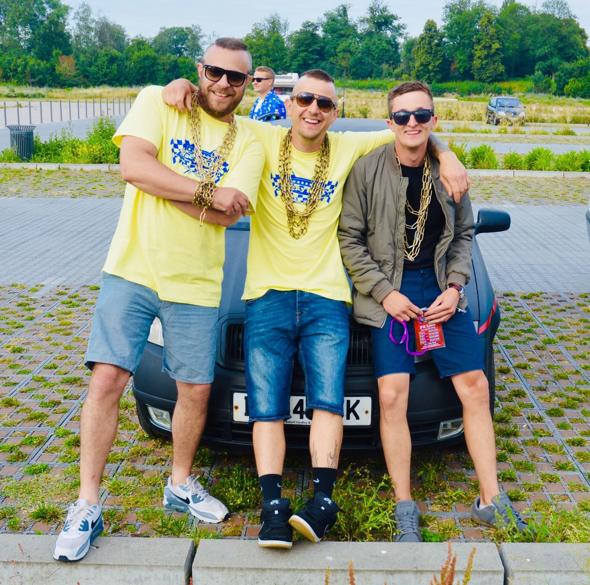 Three men standing in front of a car, wearing sunglasses and gold chains, smiling at the camera. The two on the left are wearing yellow T-shirts, while the one on the right is in a gray jacket and shorts, holding some items in his hand. They are outd