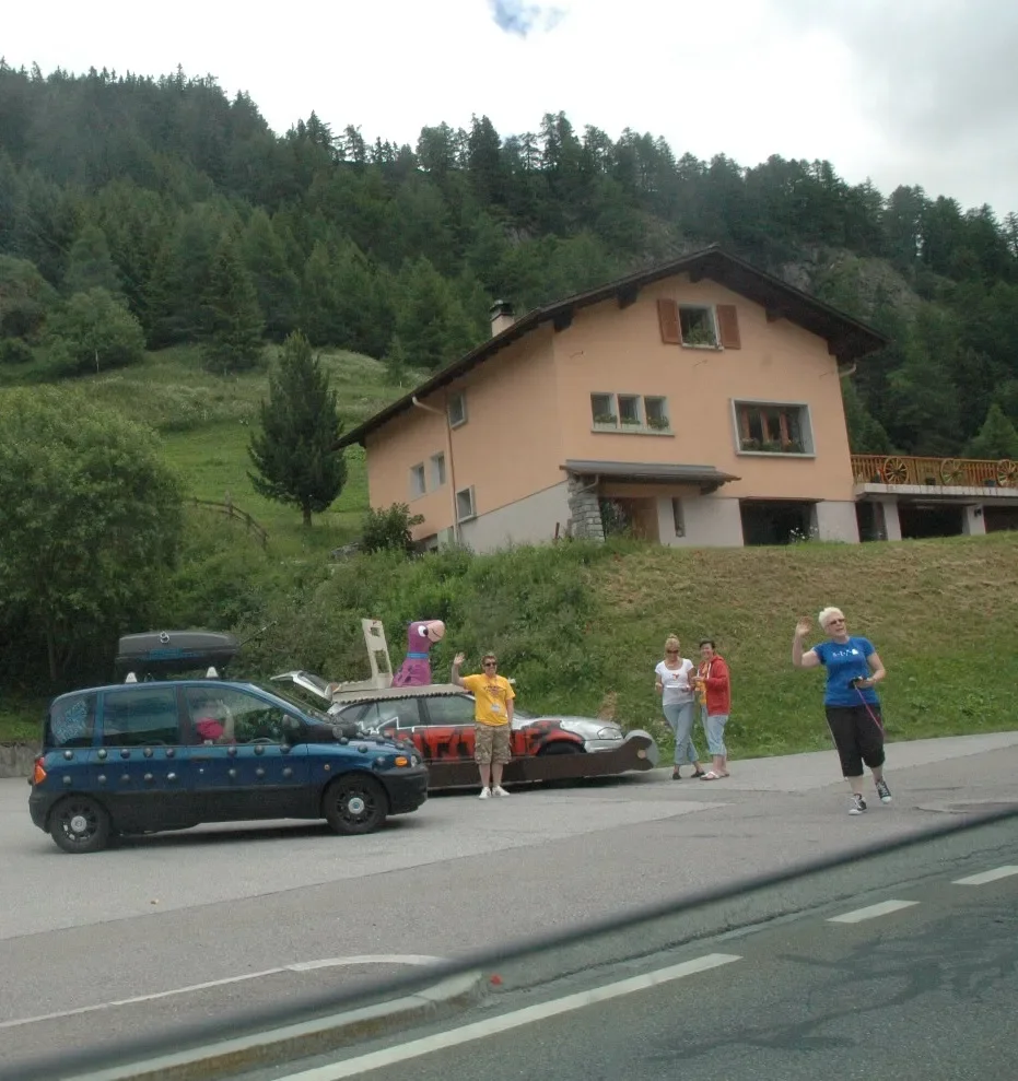 Several people standing and walking on the roadside in front of a house on a hillside; one person appears to be jogging or running; there is a parked blue car, a smaller gray car, and a pink unicorn figure near the cars. The house is large, painted l