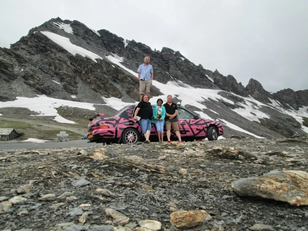 Four people posing next to a pink camouflage sports car on rocky terrain with snow-capped mountains in the background.