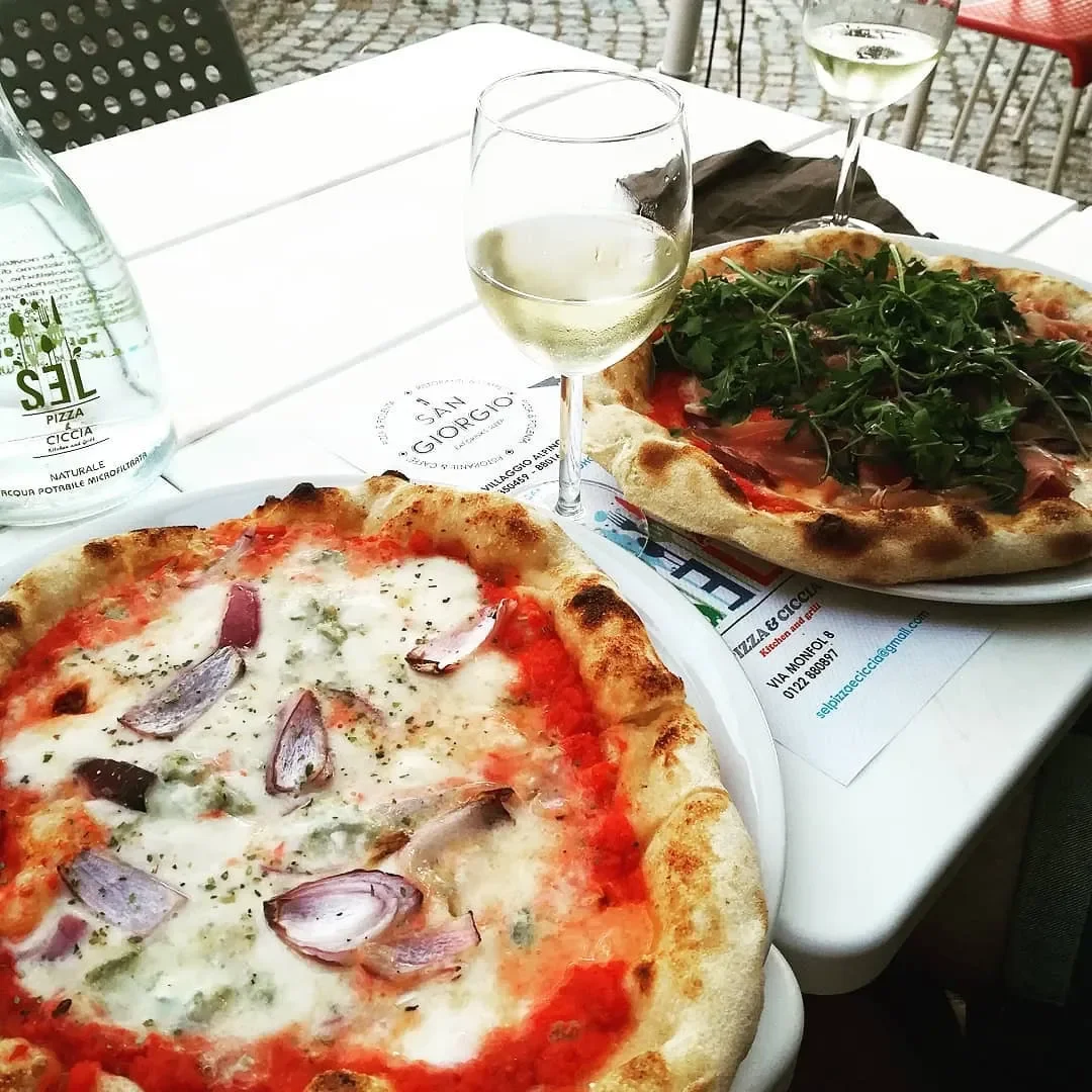 Two pizzas on white plates with glasses of white wine on a picnic table outdoors. One pizza has tomato sauce, cheese, and sliced onions. The other pizza has cheese, tomatoes, and arugula. Water bottle and black napkin are also on the table.