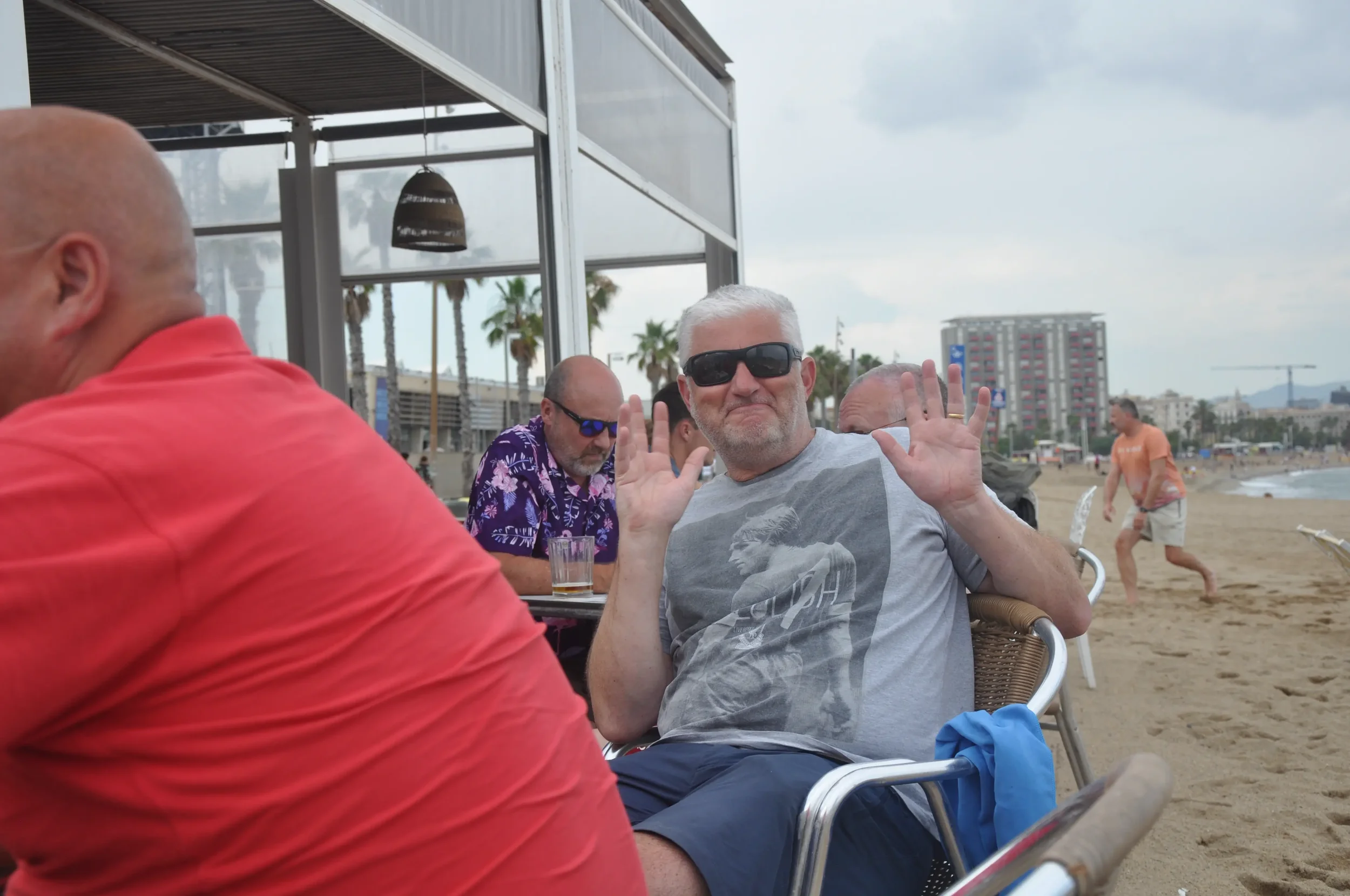 Man with white hair and beard wearing black sunglasses and a t-shirt with a graphic, sitting at a beachside cafe, waving with both hands. Other people are seated nearby, and a person is walking on the sandy beach in the background with buildings and 