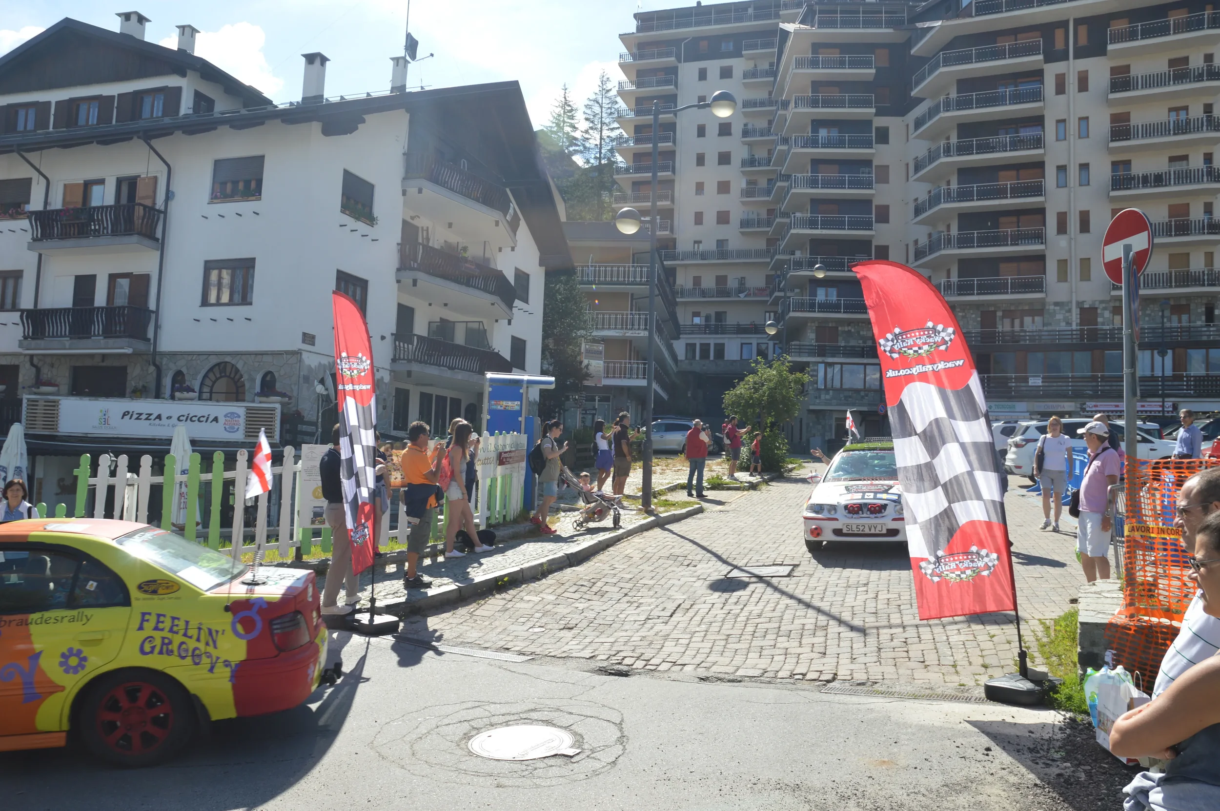 A street scene with a rally car in front of a crowd, flags, and banners at a rally event in a mountainous town with multi-story residential buildings.
