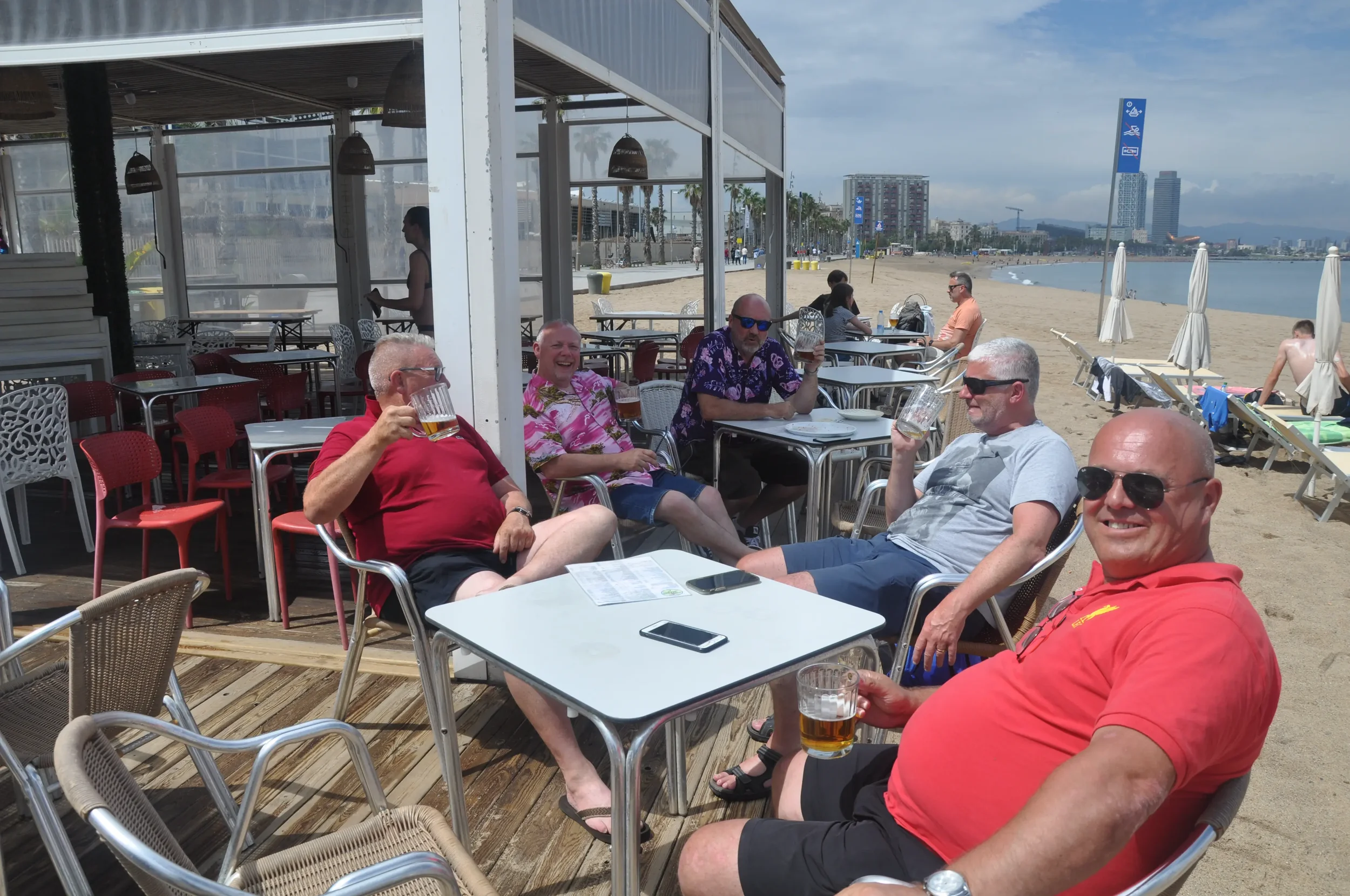 Group of five middle-aged men sitting at a table on a beachside patio with drinks, smiling, with an ocean and cityscape in the background.