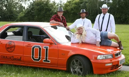A woman lying on an orange race car with the number 01, surrounded by three men in cowboy hats, in a grassy field.