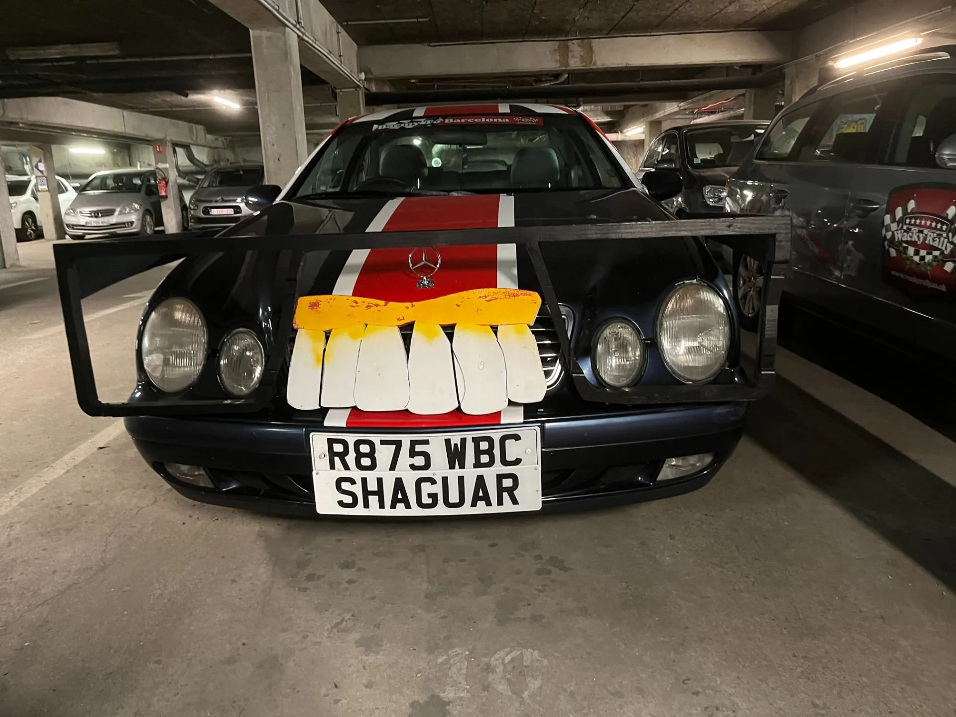 A black car with red racing stripes and a Mercedes-Benz logo parked in a multi-level parking garage. The car has a black frame with a white, yellow, and orange decorative piece attached to the front grill, resembling a face with large eyes and a yell
