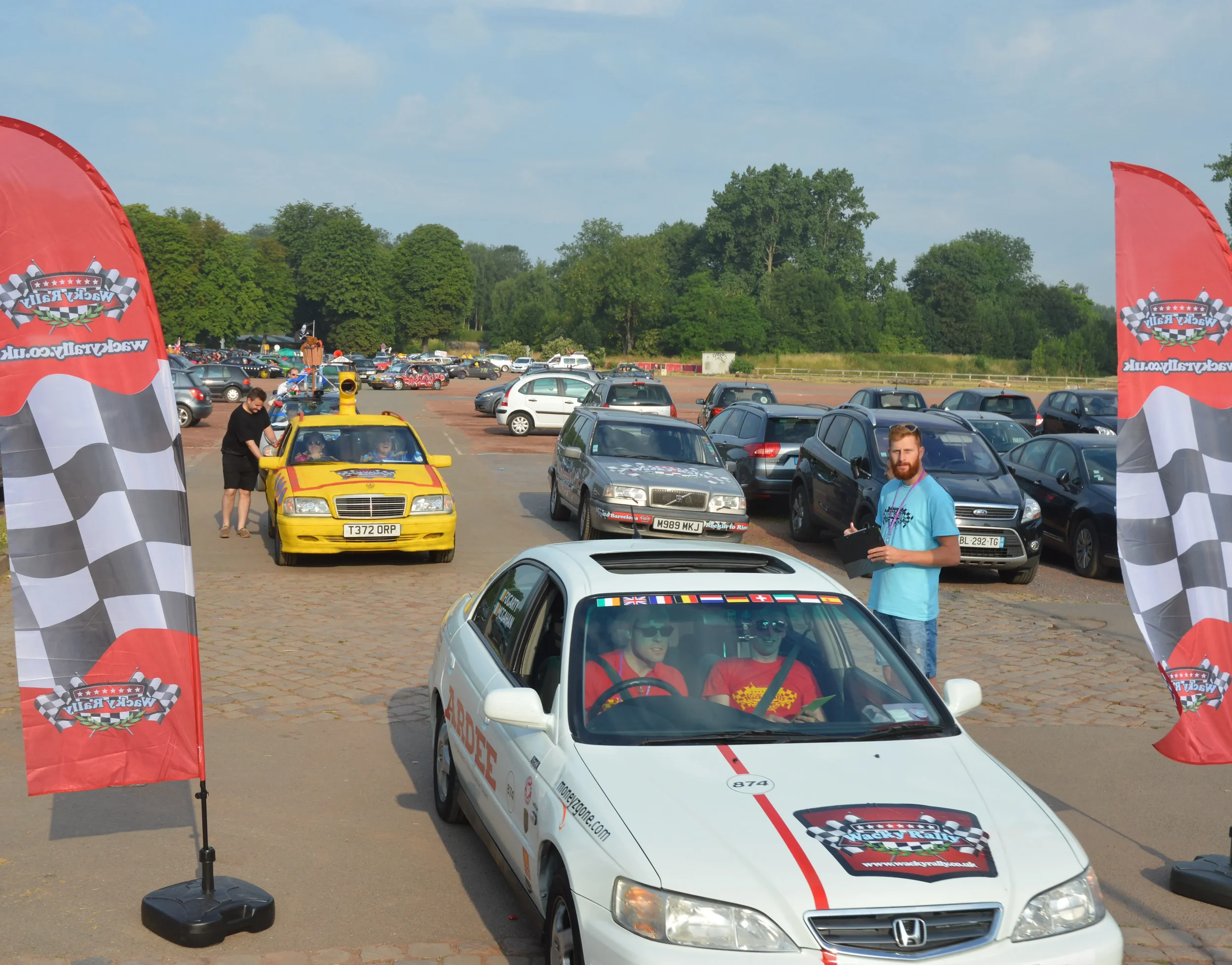 A white Honda car at a race event with a rally logo, cars and parking lot in the background, two flags with checkered pattern and rally logo on each side, a man with a clipboard, and other vehicles including a yellow car with a rally-themed paint job