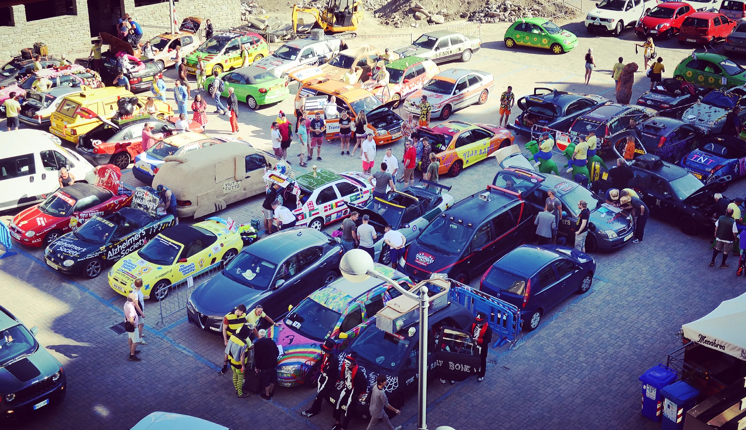 A lively outdoor car event featuring a variety of decorated and customized cars, with people walking around, chatting, and viewing the vehicles. Some cars have costumes or themed decorations, and there are inflatables and costumes among the attendees