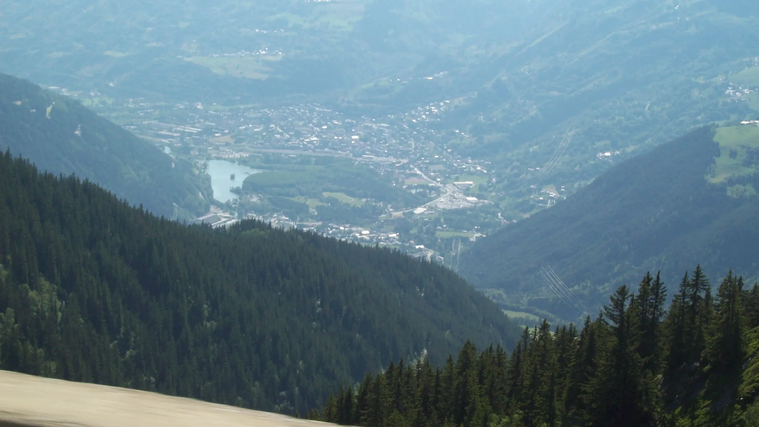 Aerial view of a mountain valley with a river, surrounded by dense forests and small town buildings, under a hazy sky.