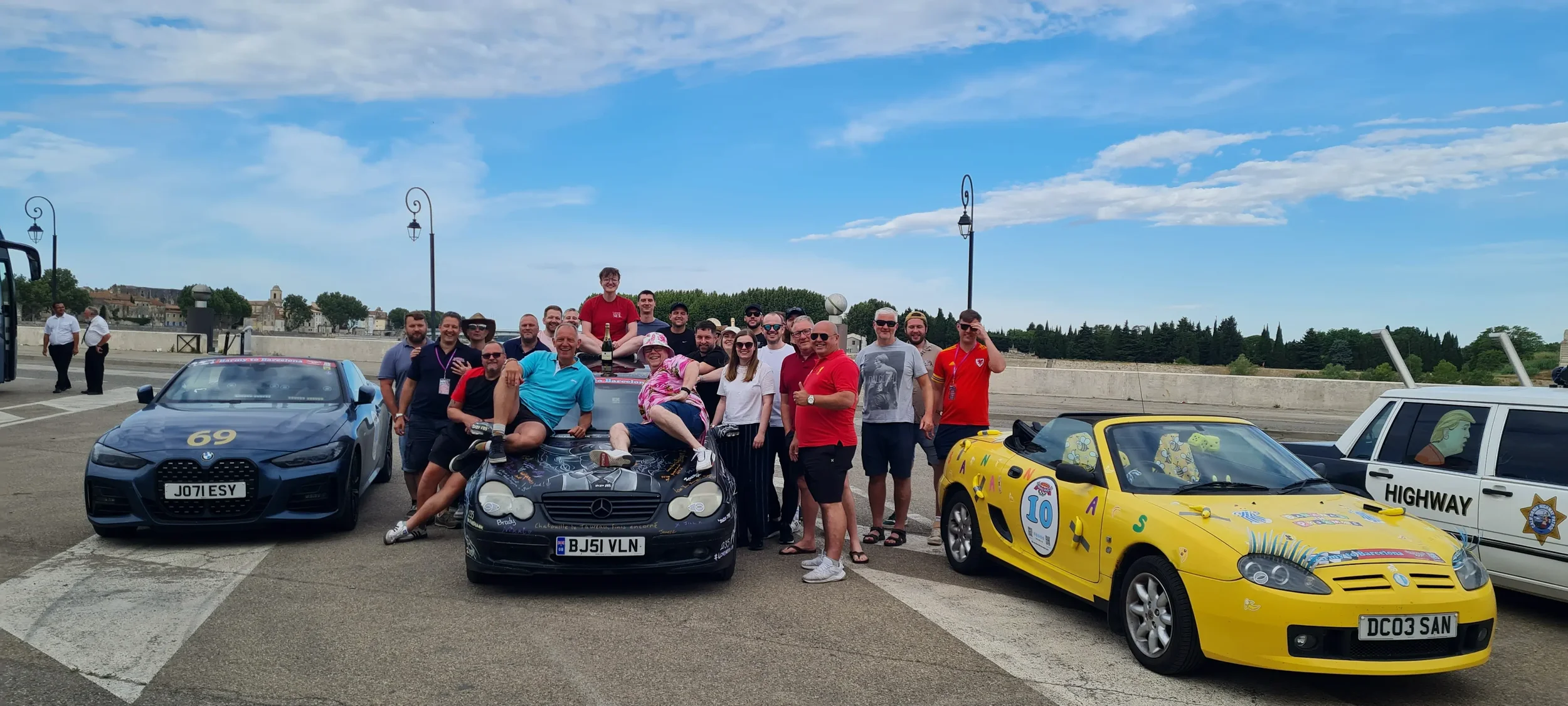 Group of people gathered around three cars on a rooftop parking lot with a scenic view and a blue sky.