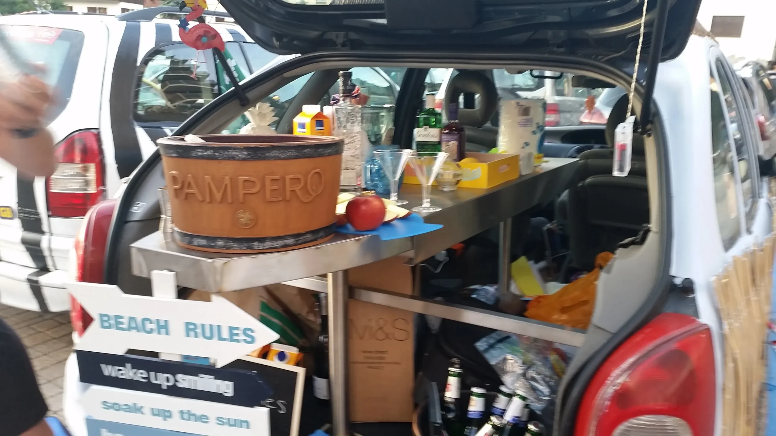 Open vehicle trunk with a table holding drinks, snacks, bottles, and an apple, at an outdoor market or event.