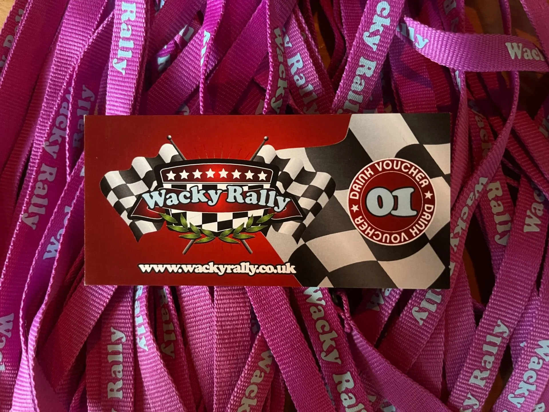 A Wacky Rally event card with a racing flag theme and a pink wristband with 'Wacky Rally' written on it. The card features a red and black checkered racing motif, a logo with checkered flags, and the website www.wackyrally.co.uk.