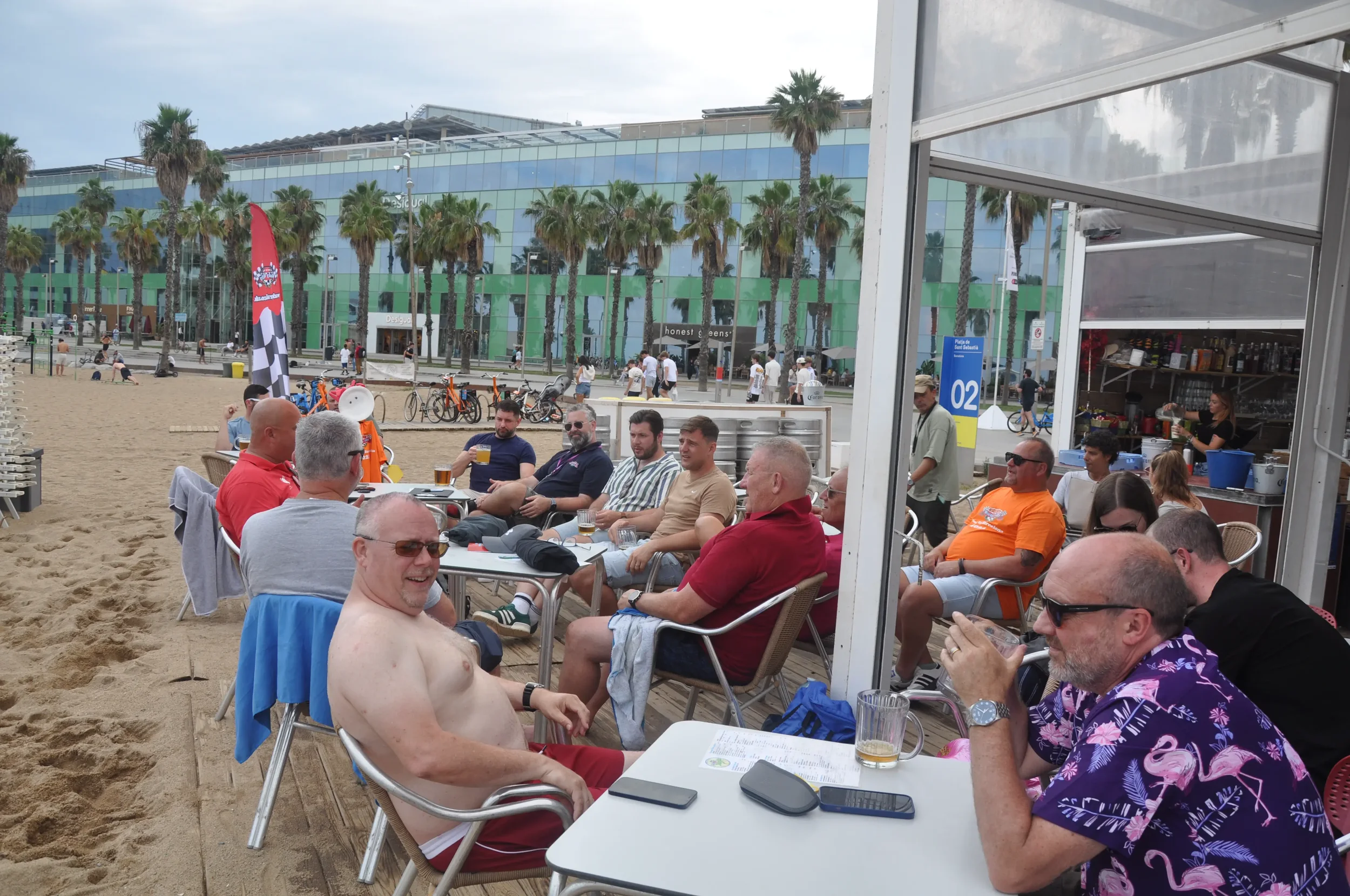 Group of people sitting at outdoor tables on a beach boardwalk, some with drinks, in front of a beachside bar with palm trees and a modern building in the background.