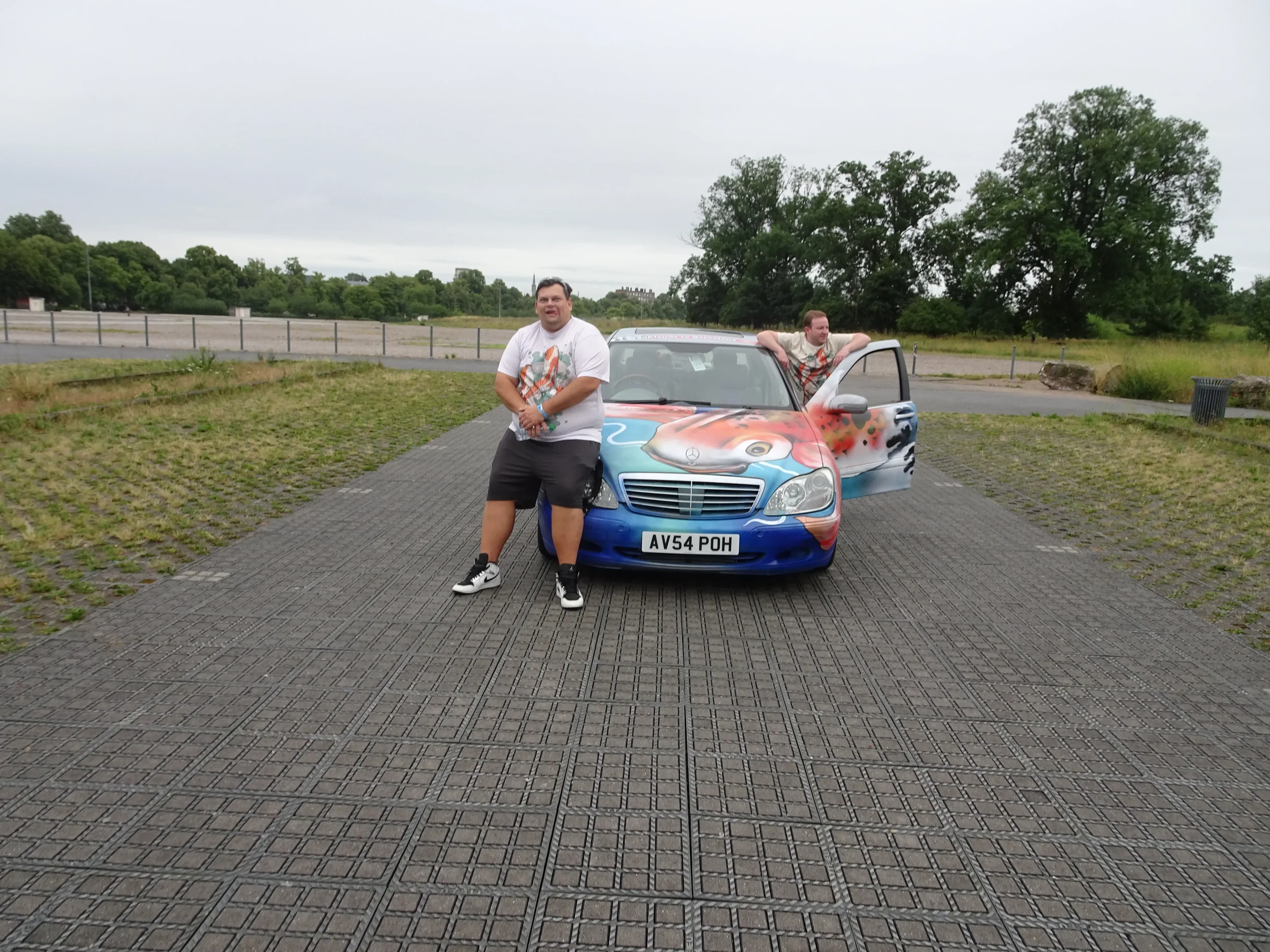 Two men with a colorful painted car, one standing in front and one sitting in the car's open door, in an outdoor parking area with grass, trees, and cloudy sky.
