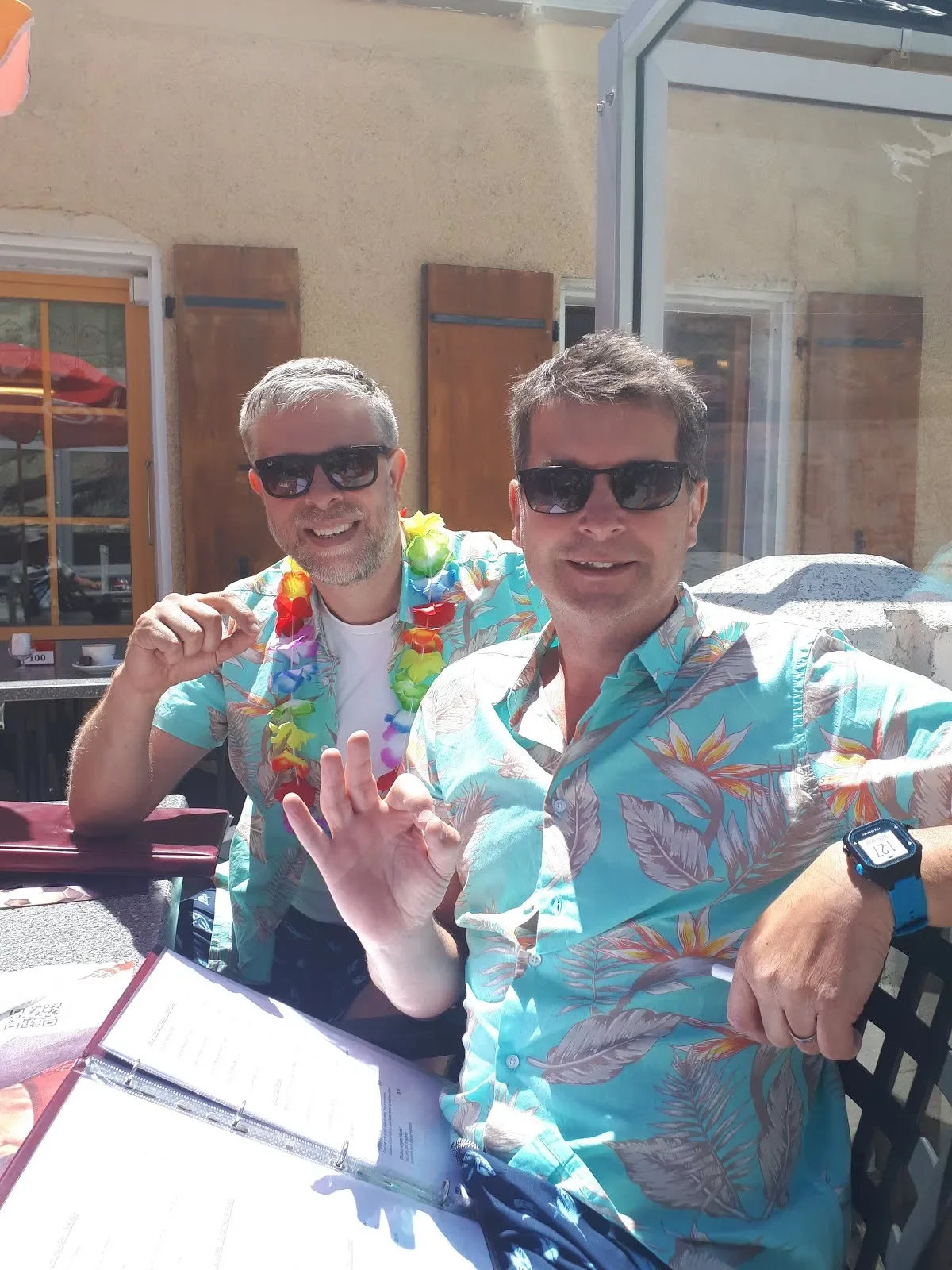 Two men wearing tropical shirts and sunglasses sitting at an outdoor table, one making an OK gesture, celebrating a summer or vacation occasion.