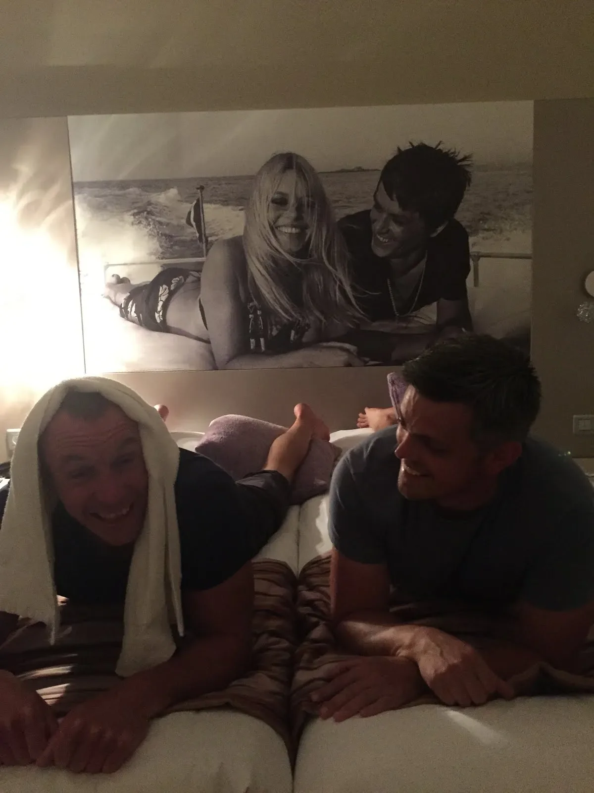 Two men lying on bed, one with a towel over head, smiling and looking at each other, with a large black-and-white photograph of a woman and man on the wall behind them.