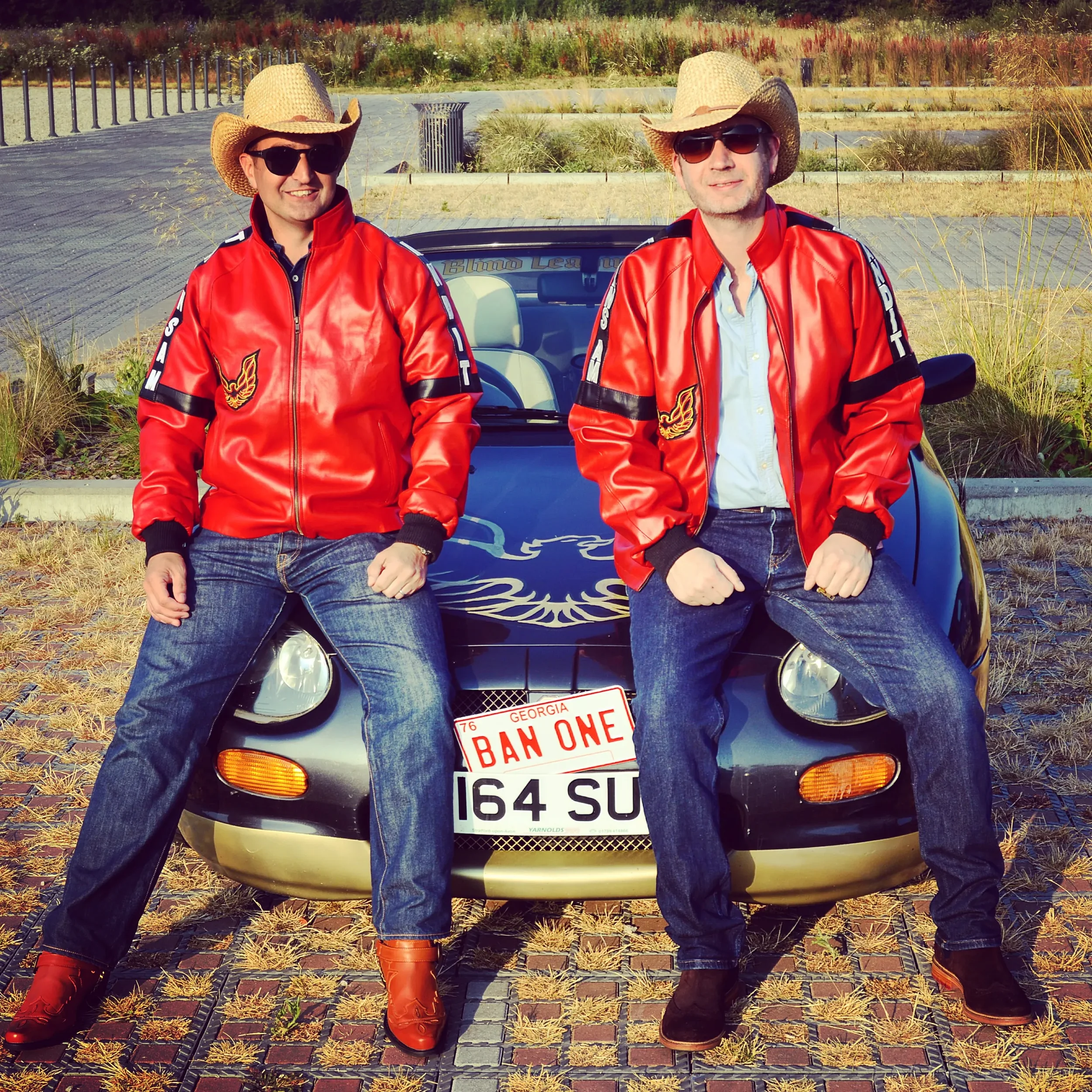 Two men wearing red leather jackets, cowboy hats, sunglasses, and jeans, sitting in front of a black sports car with Georgia license plates, in a parking lot surrounded by plants and landscaping.