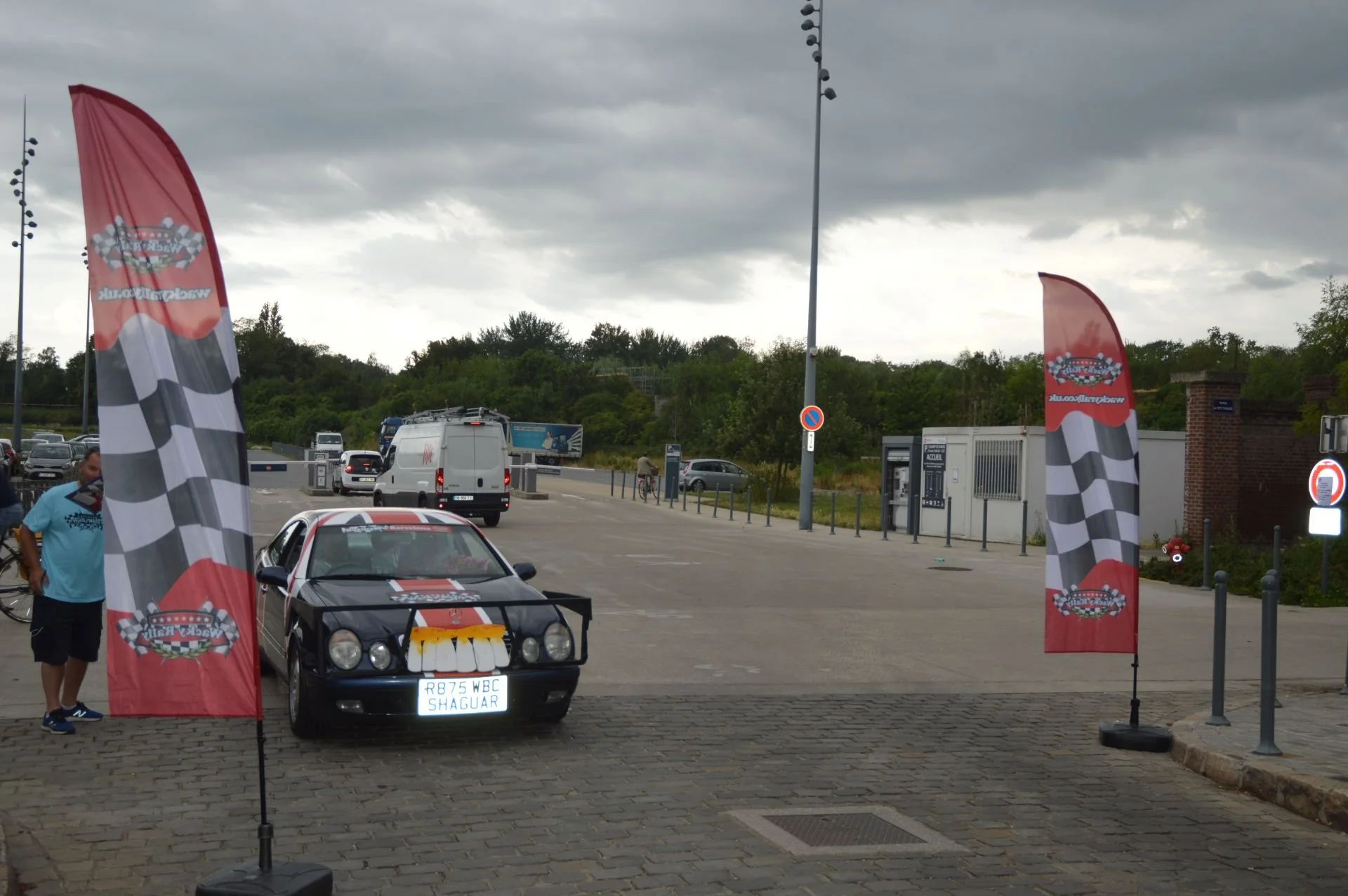 A black classic car with racing decals and a license plate reading 'R875 WBC SHAGUAR' parked between two tall checkered flags at a car event, with other vehicles and people in the background under a cloudy sky.
