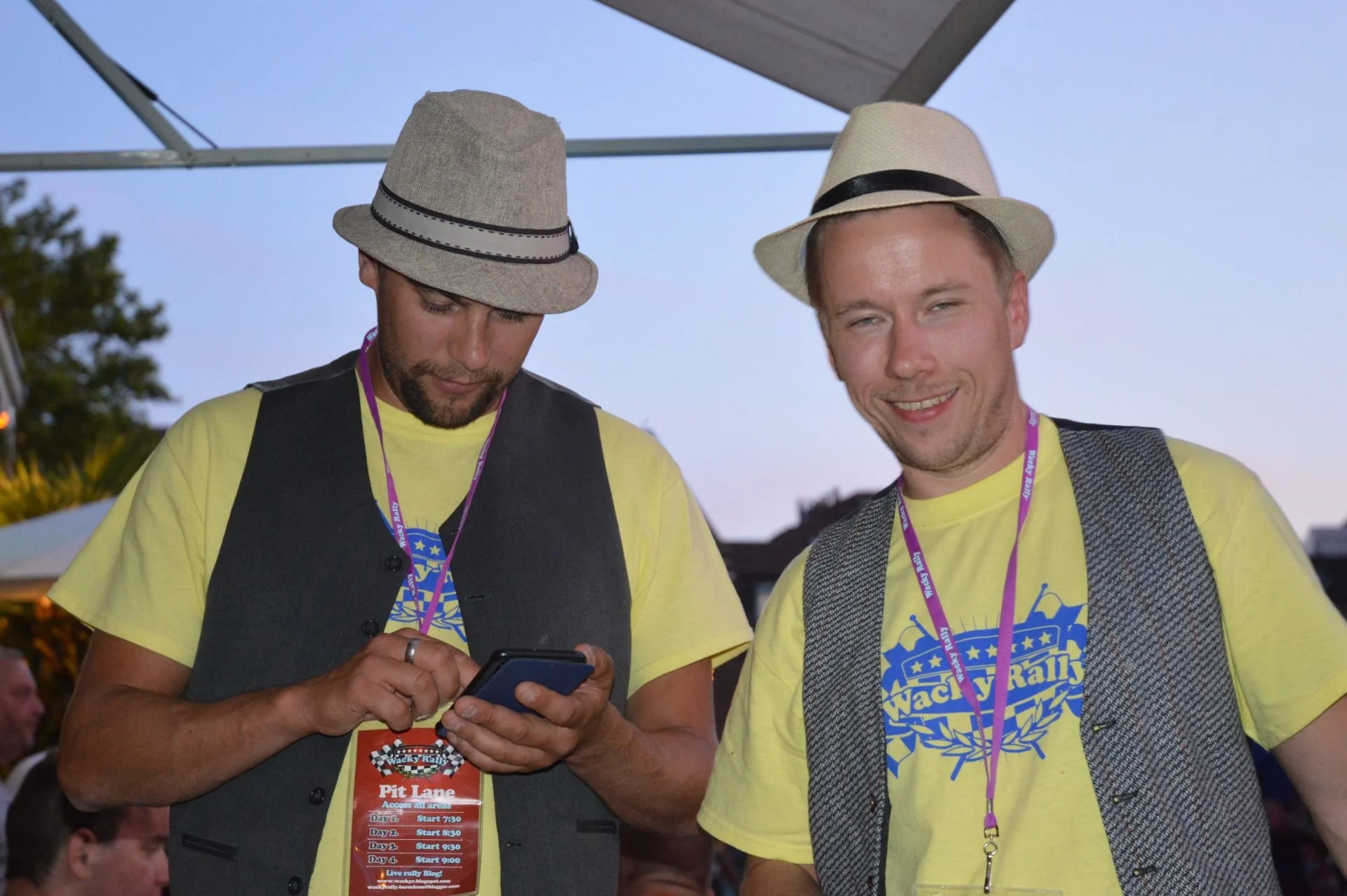 Two men at an outdoor event wearing yellow t-shirts and beige hats. One man is looking at his phone, the other is smiling at the camera. They are wearing event badges on lanyards around their necks.